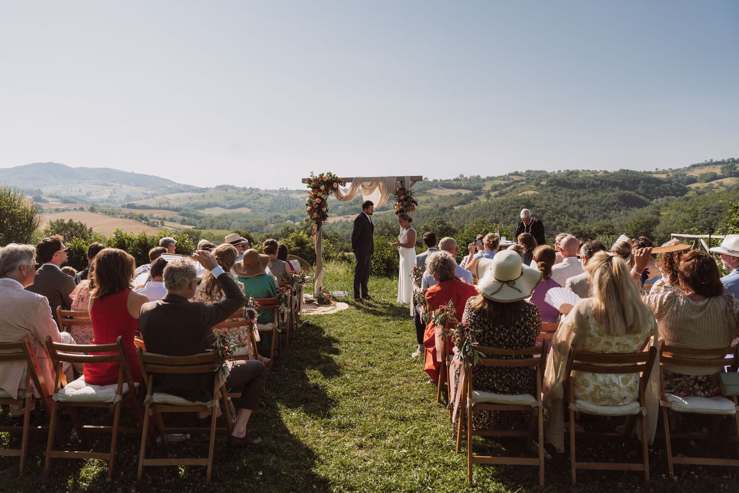 Guests seated in wooden chairs at an outdoor wedding ceremony overlooking scenic mountain views in the countryside.