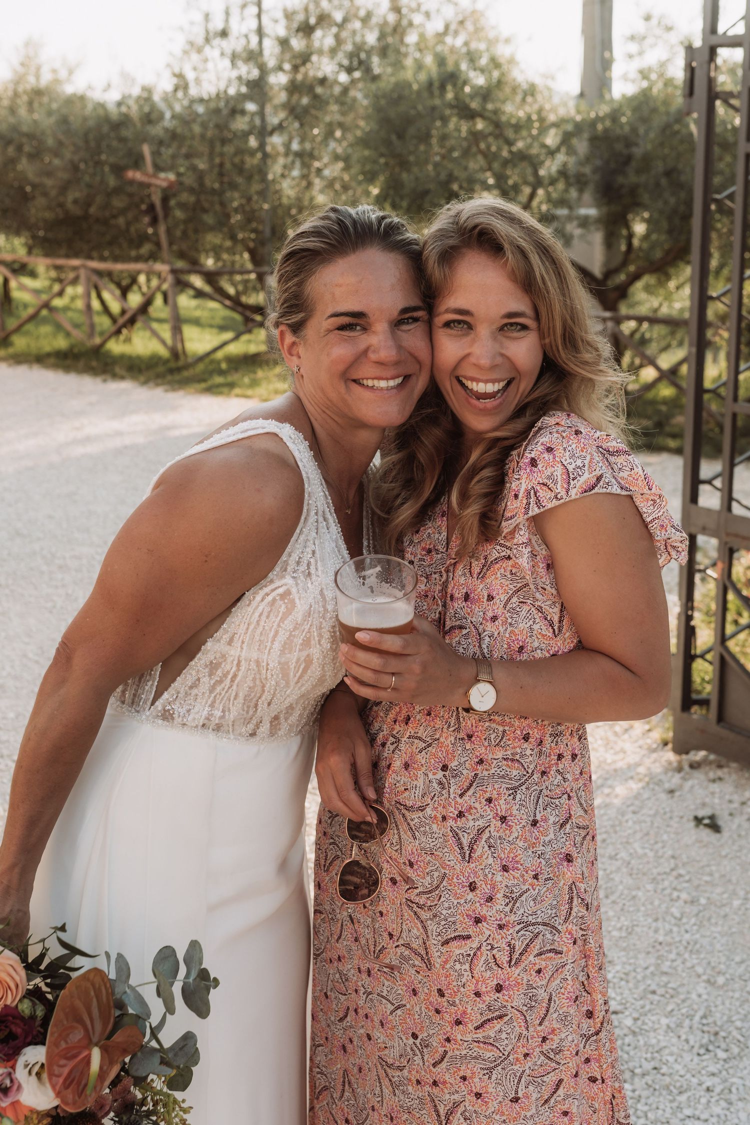 Two people in summer dresses share a happy moment outdoors while holding drinks and flowers on a sunny day.