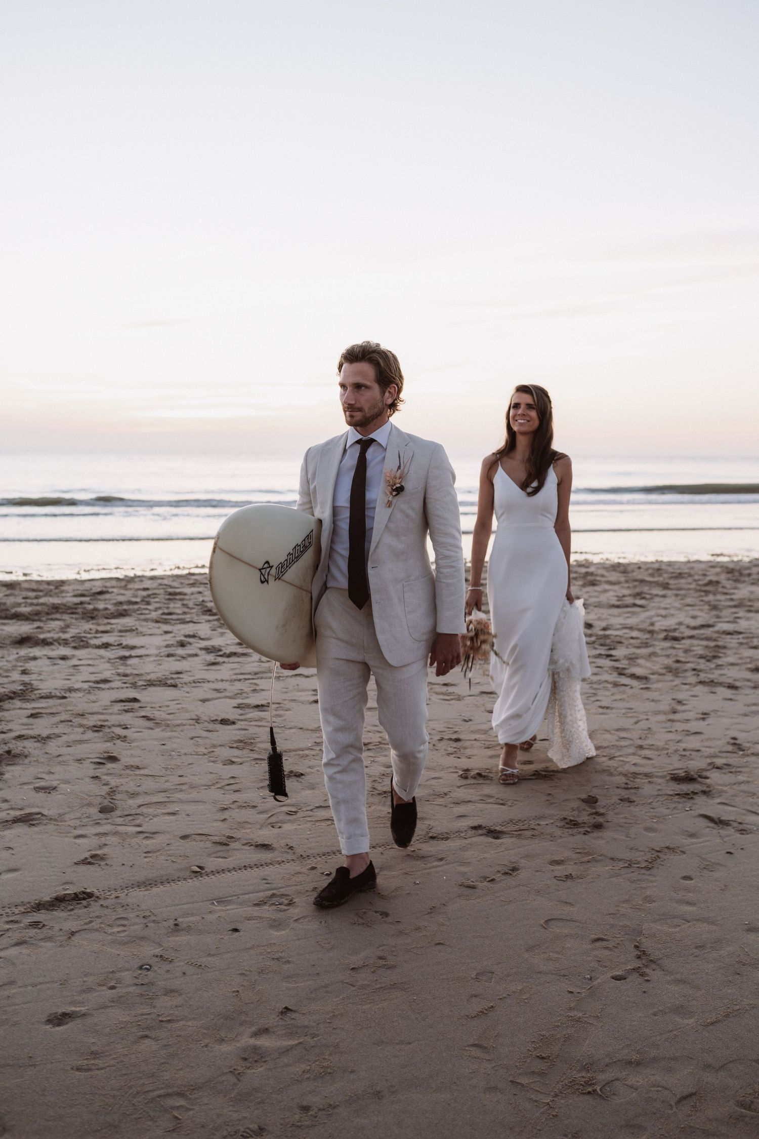A couple in white attire walking along a beach at sunset with a surfboard.