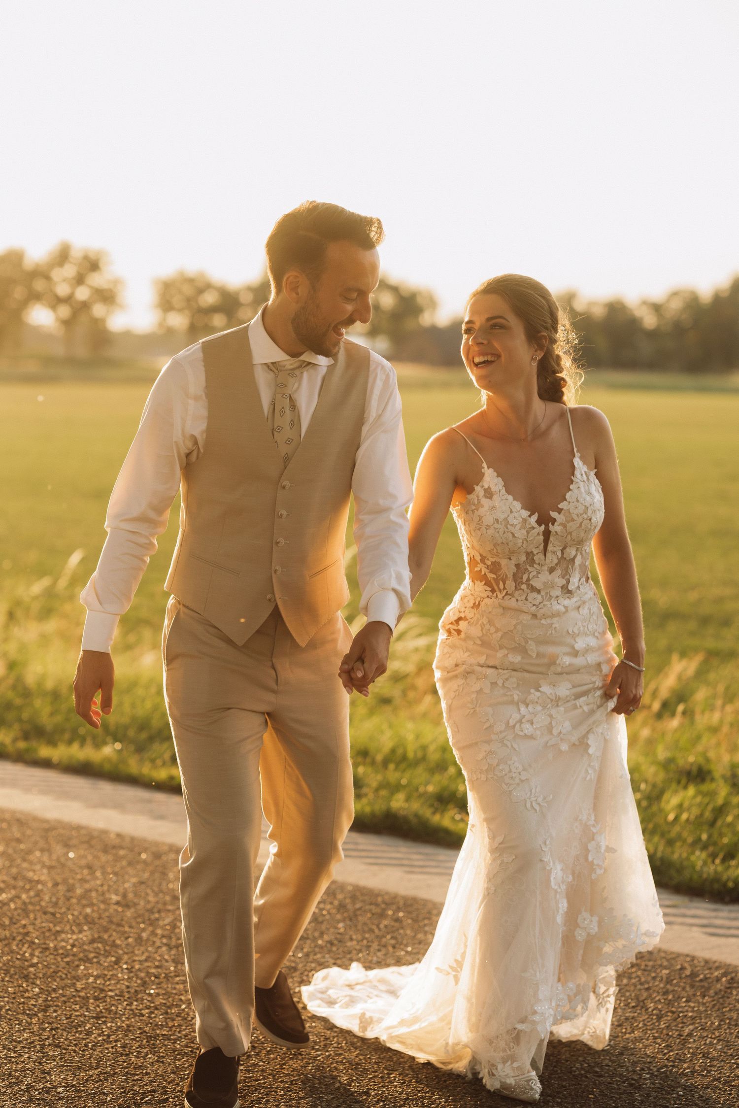 Newlyweds walk together on a path during sunset with the bride in a detailed lace dress.