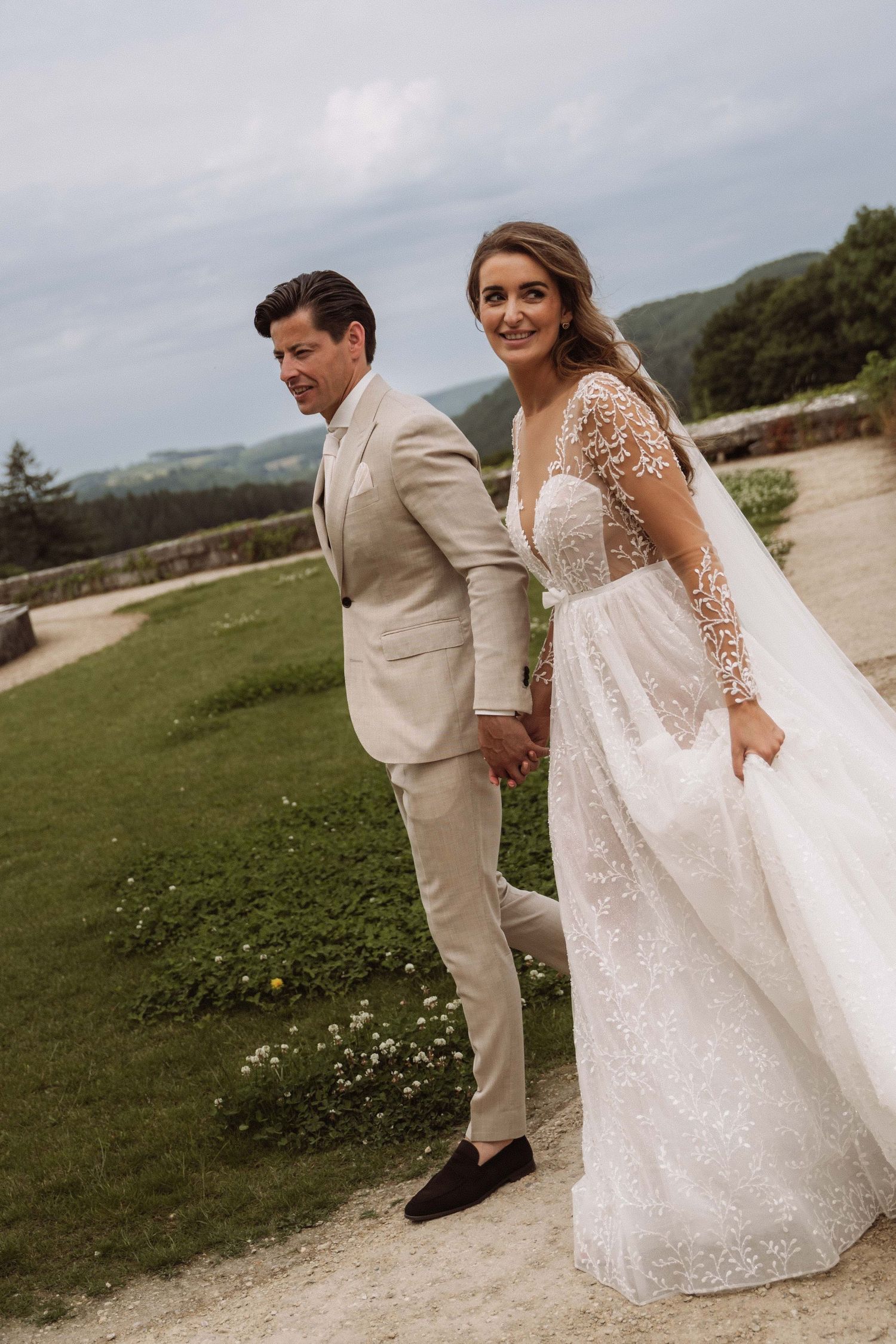 Romantic wedding couple poses outdoors on a grassy hillside path with flowing white dress and beige suit.