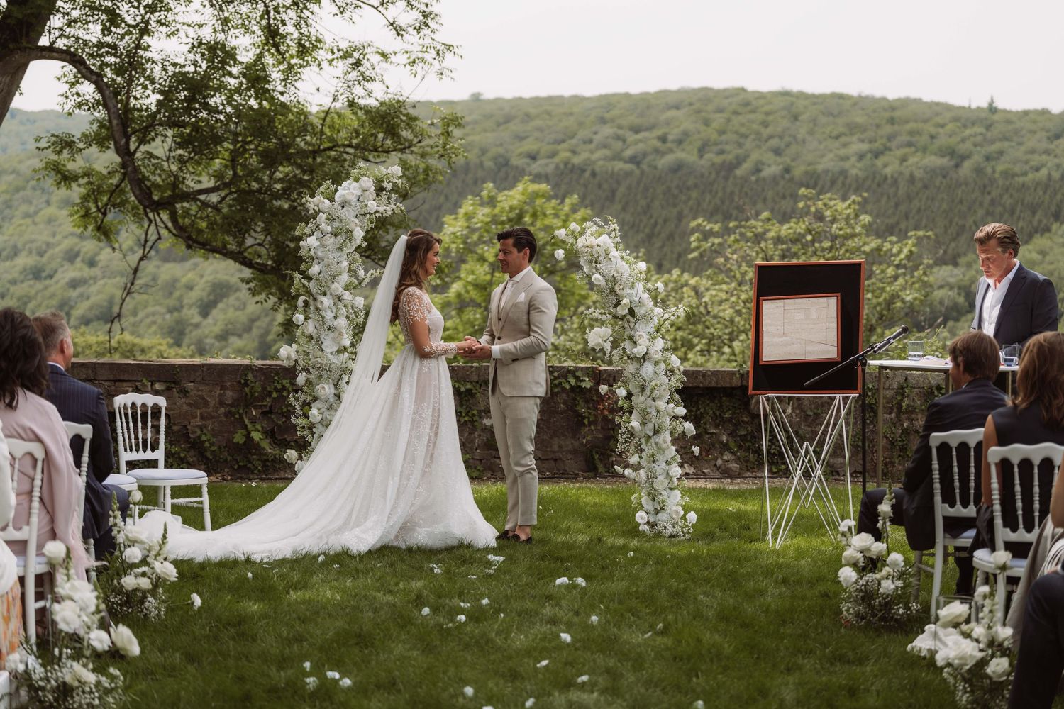 An outdoor wedding ceremony set against rolling green hills with white chairs and floral arrangements creating an elegant aisle.