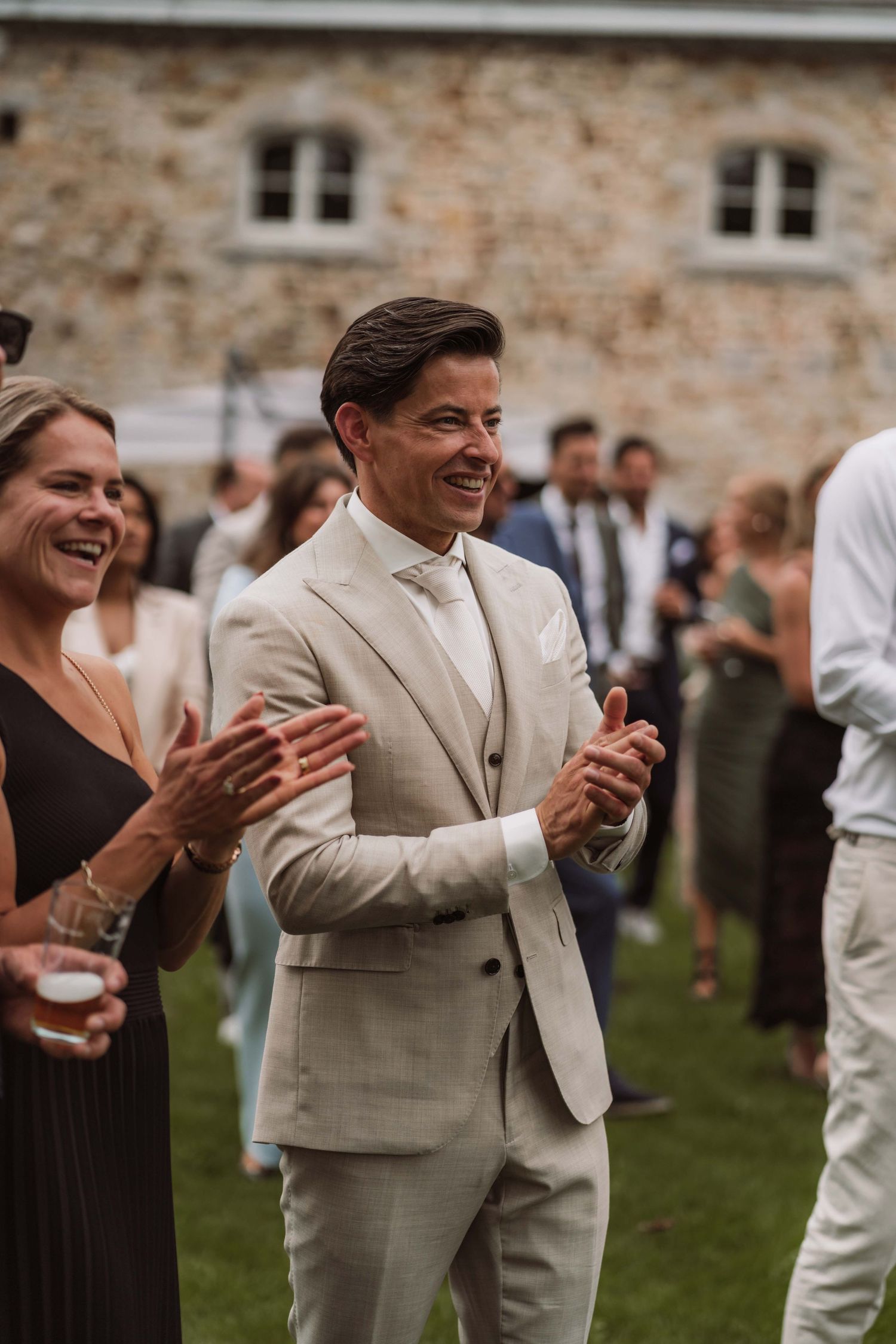 Wedding guests in beige and dark attire share joyful laughter during an outdoor celebration on a lawn.