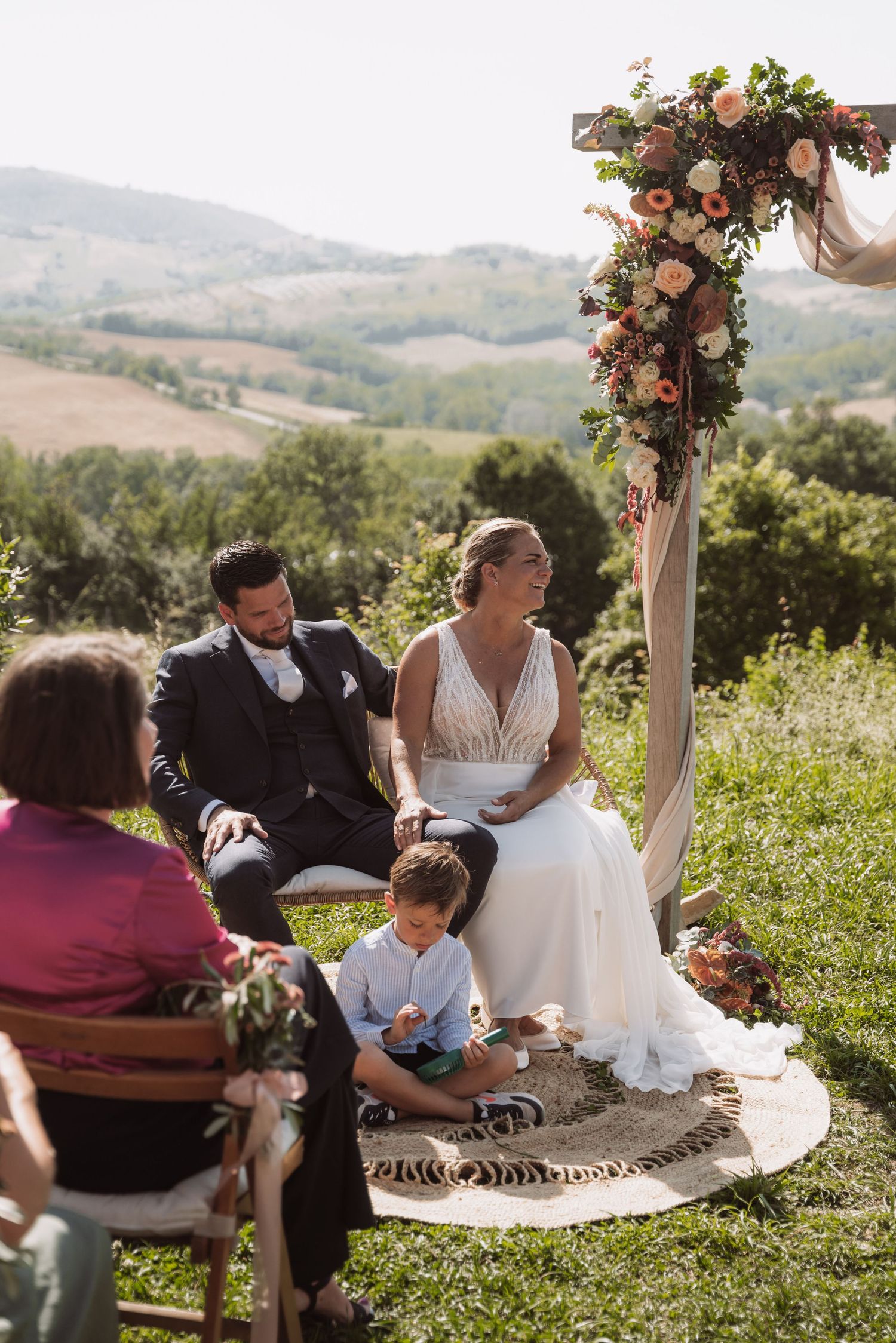Wedding party relaxes on a blanket during outdoor hillside celebration with floral arch decorations in scenic setting.