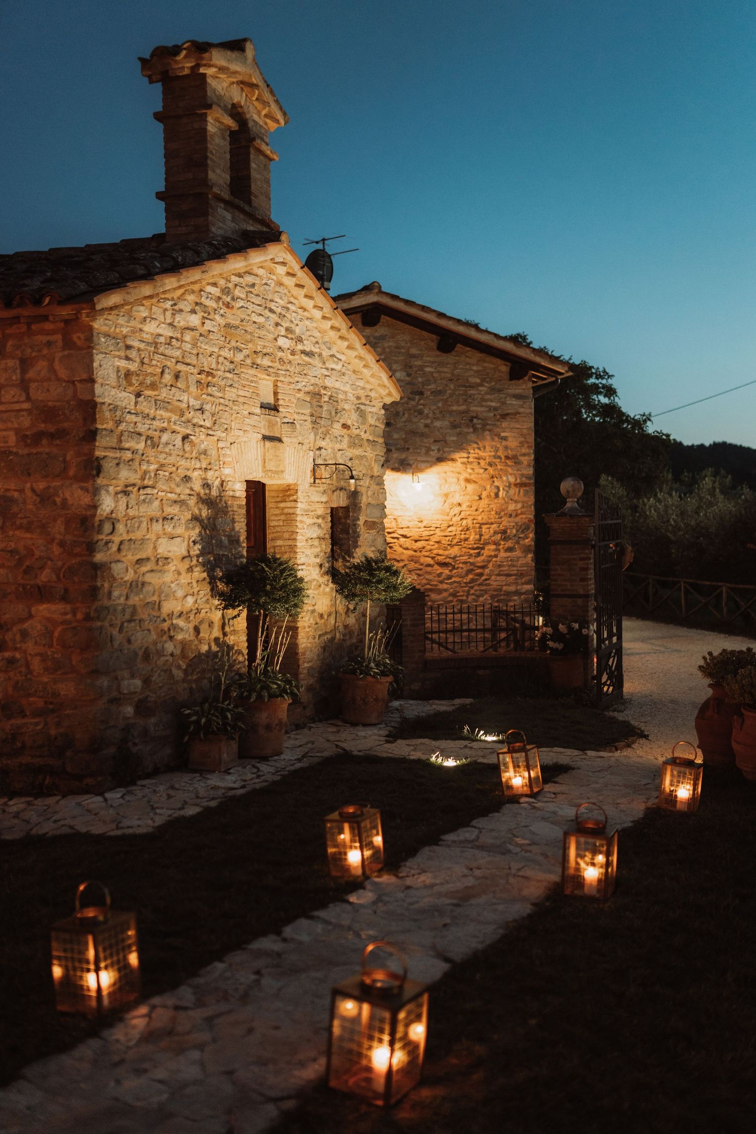 Stone cottage pathway illuminated by lanterns at dusk with warm glowing lights against blue sky.