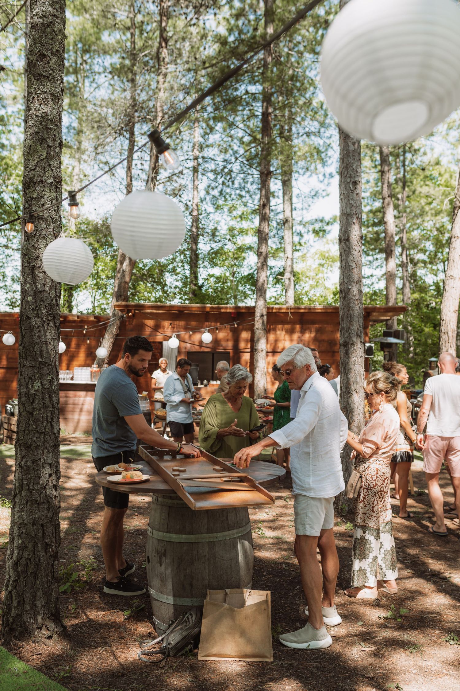People gather around wooden barrels in a pine forest setting decorated with white paper lanterns for an outdoor event.