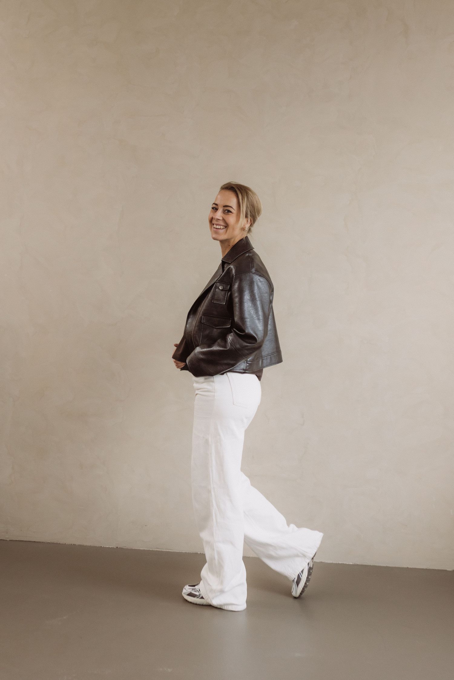 A figure in a leather jacket and white pants strikes dynamic poses against a neutral beige wall in a photo series.