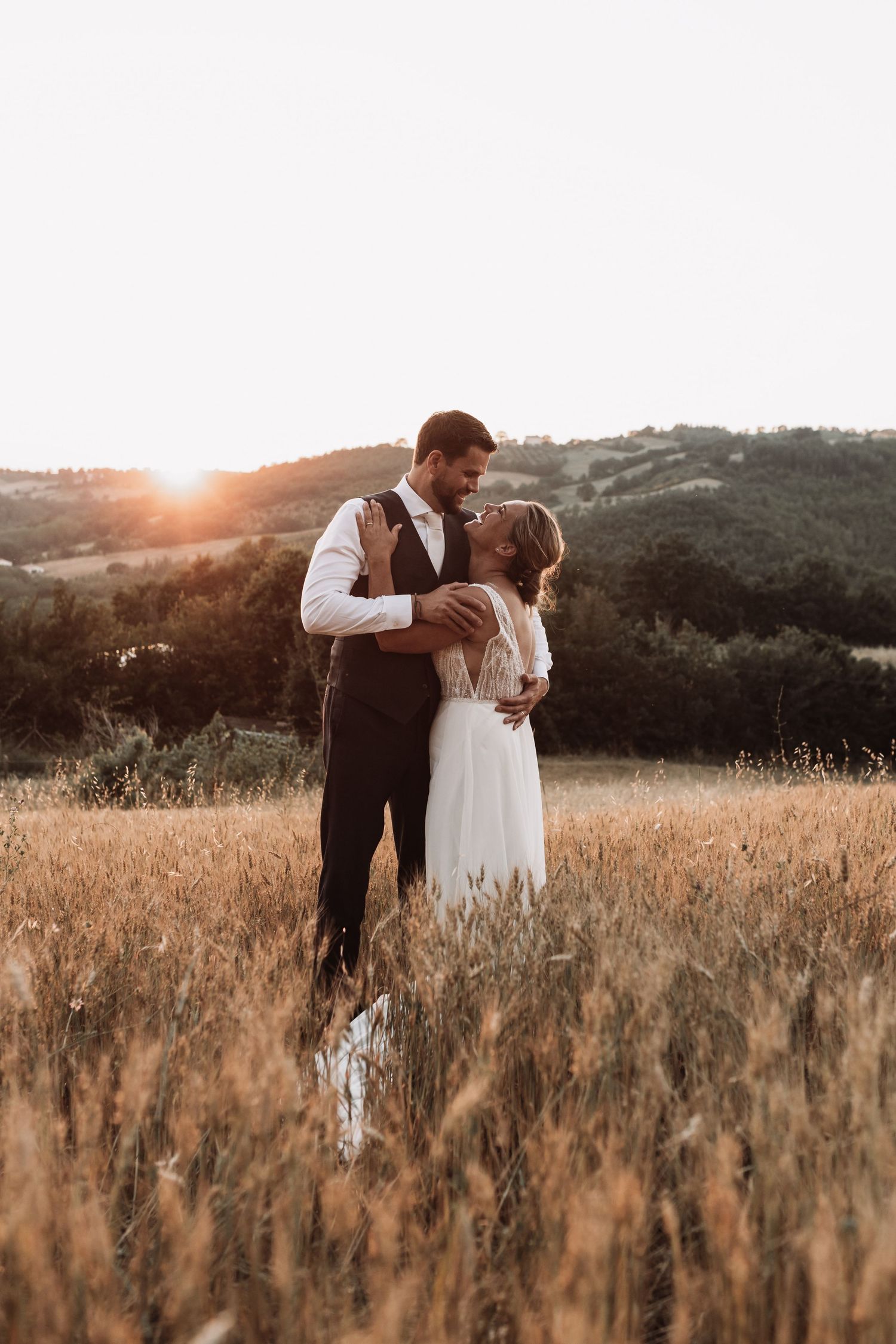 A couple in wedding attire embraces in a golden field at sunset with mountains in the background.