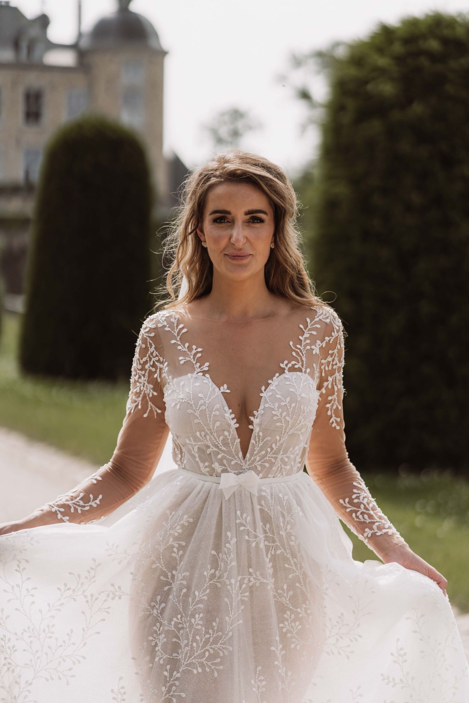 A romantic bridal gown with delicate lace details and flowing tulle skirt shown in an outdoor garden setting.