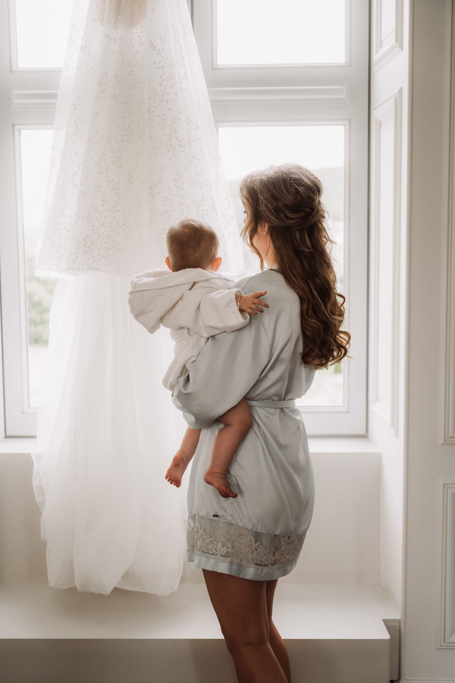 Child in grey sweater plays with white curtains near bright window creating dreamy atmosphere.