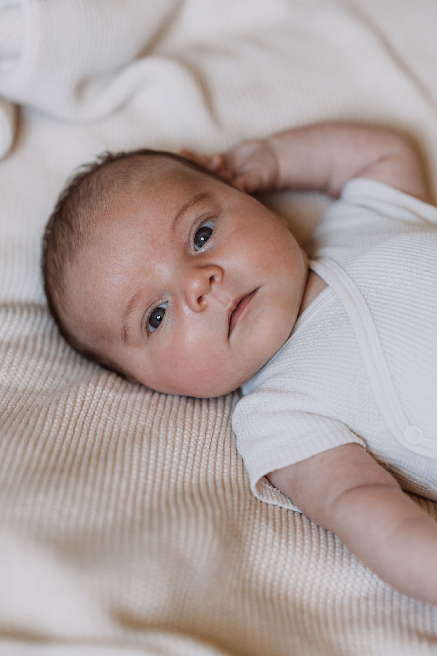 Baby in white onesie lying on textured fabric looking up with bright eyes.