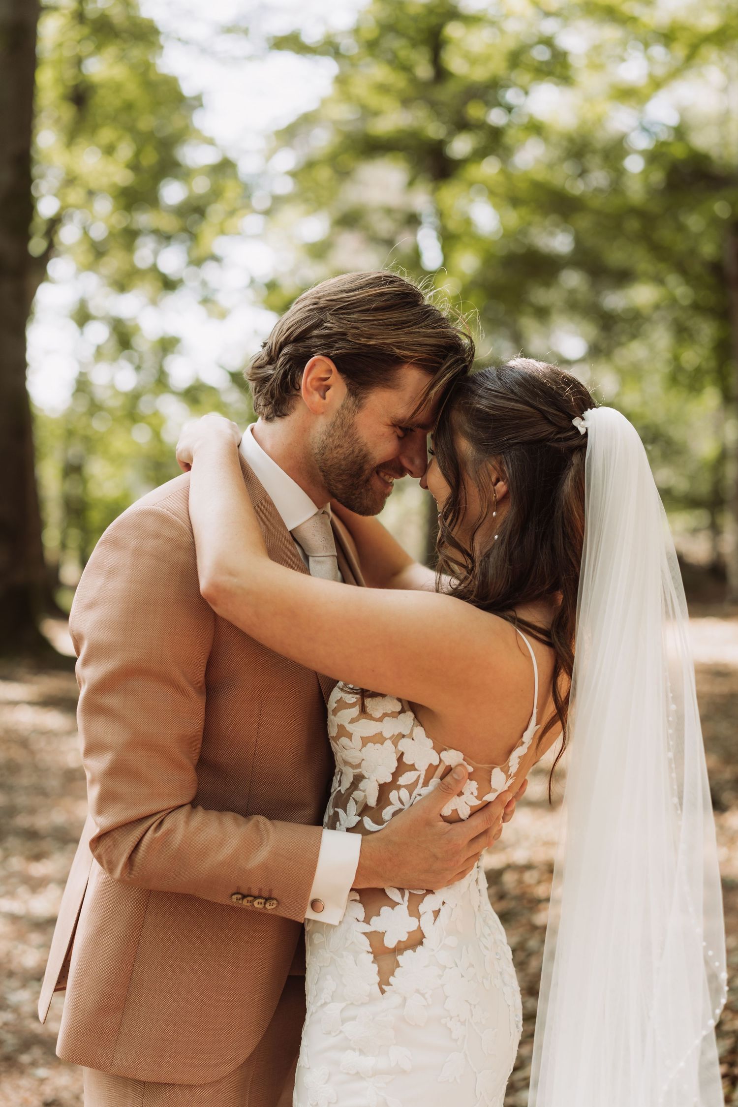 Romantic wedding couple embraces in a sunlit forest setting, bride wearing lace dress with veil and groom in tan suit.
