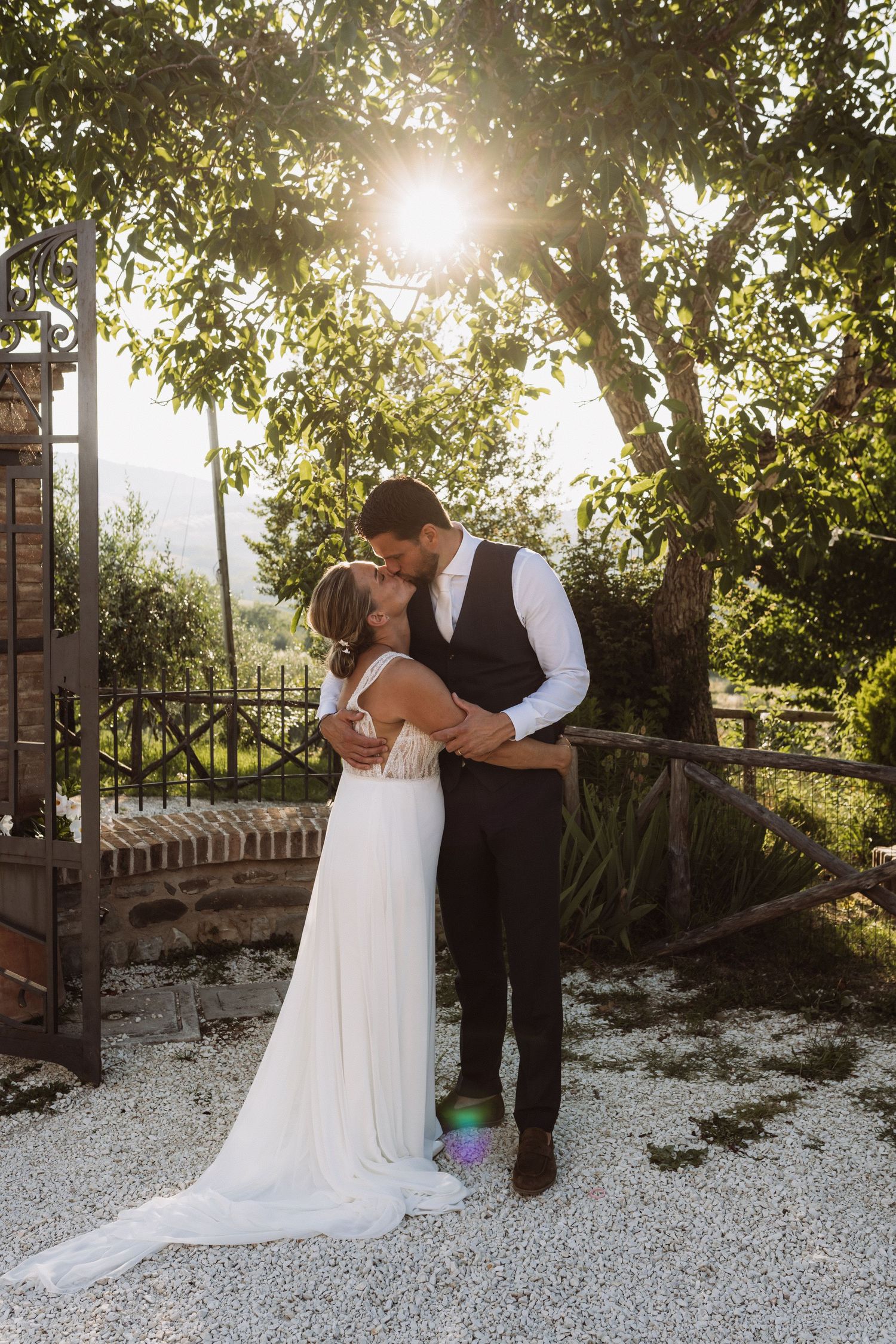 A romantic wedding couple shares tender moments in a sunlit garden with white gravel and lush greenery.