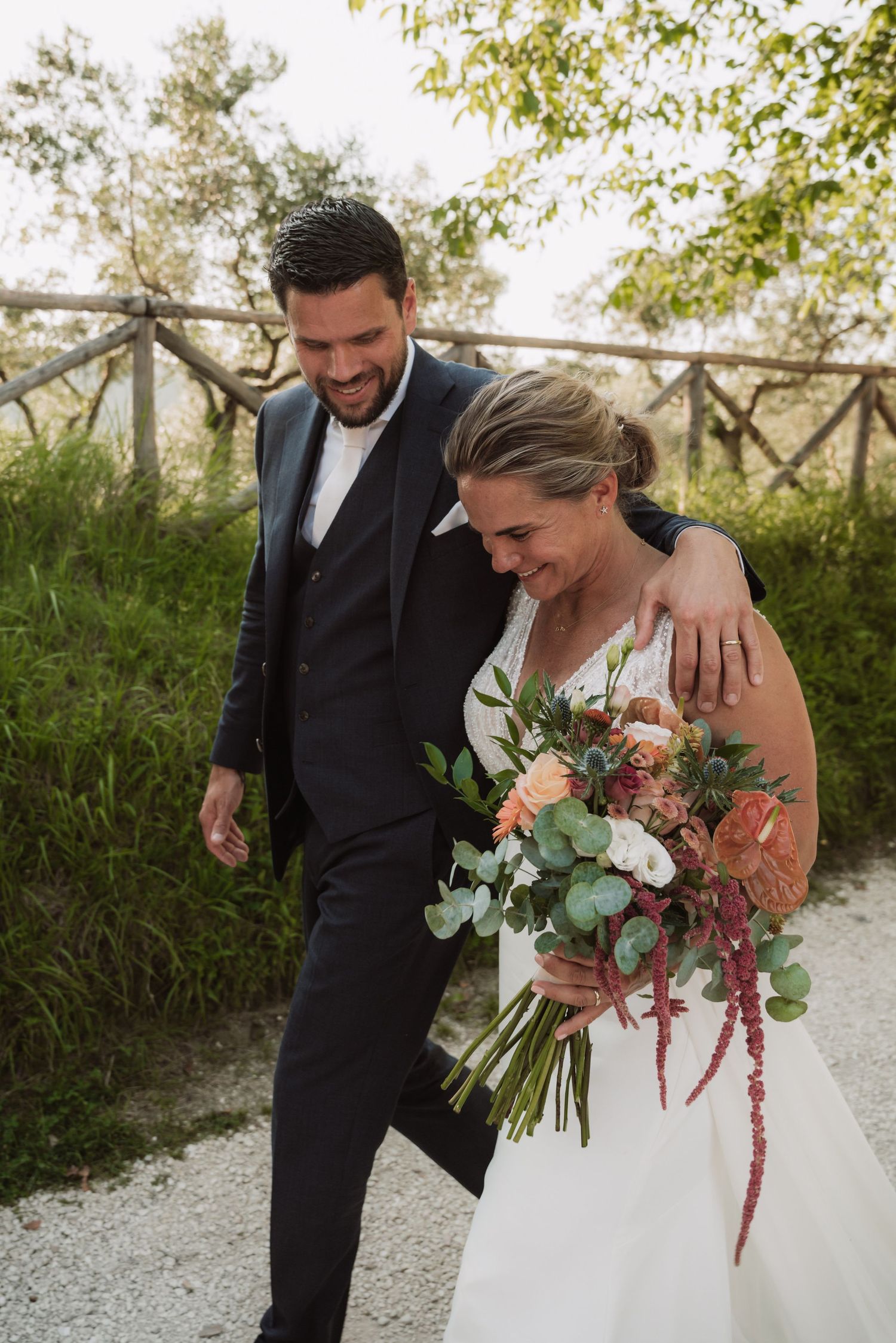 A joyful couple walks together on a gravel path, with the bride holding a beautiful bouquet of pink and white flowers.