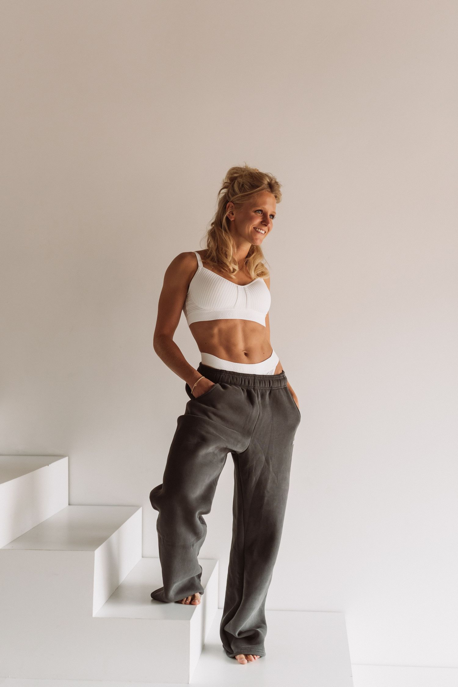 Woman in white crop top and gray pants poses against white wall in minimalist fashion photo series.