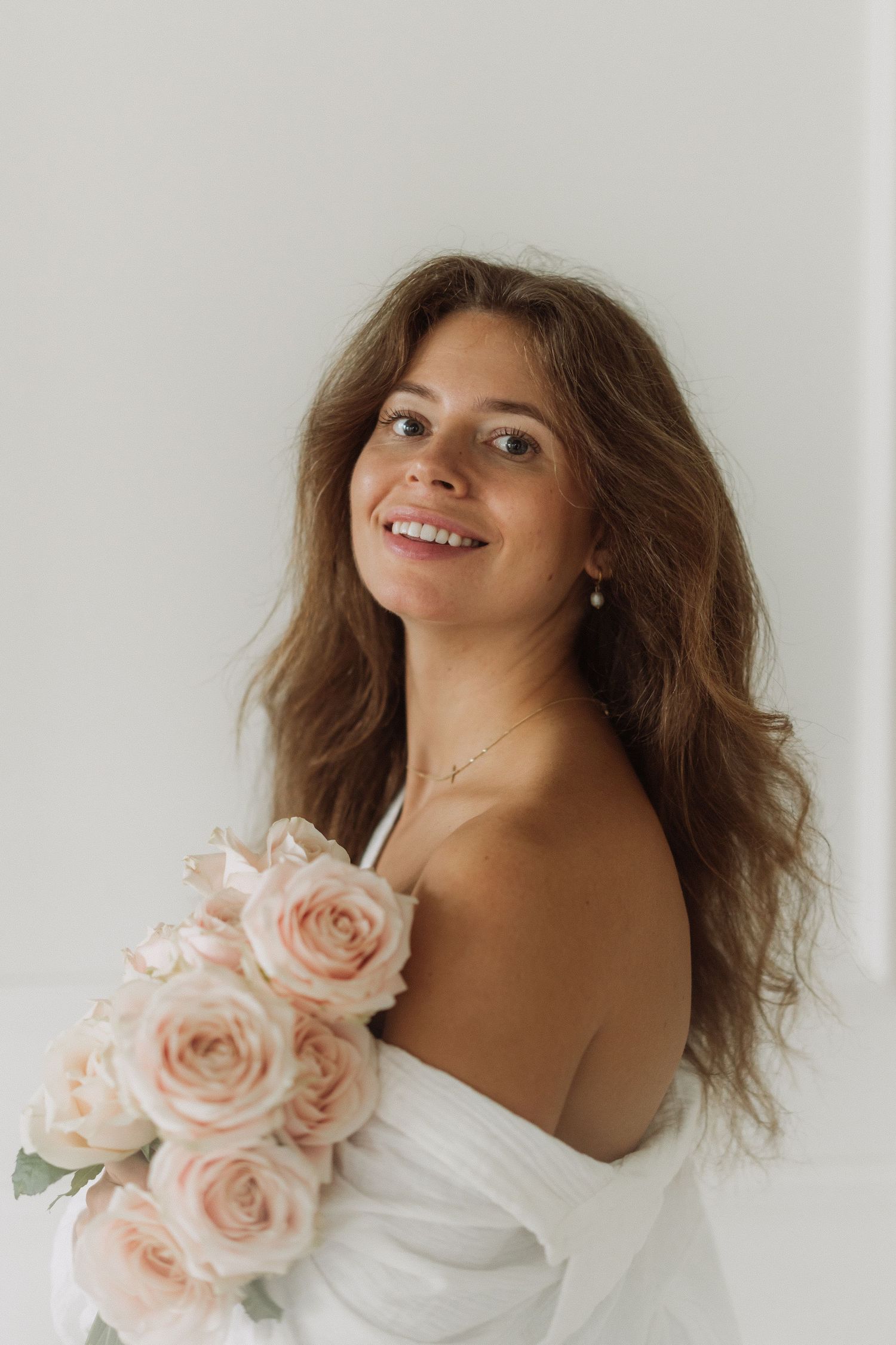 Woman with wavy hair holds pink roses while posing against white wall in romantic portrait series.