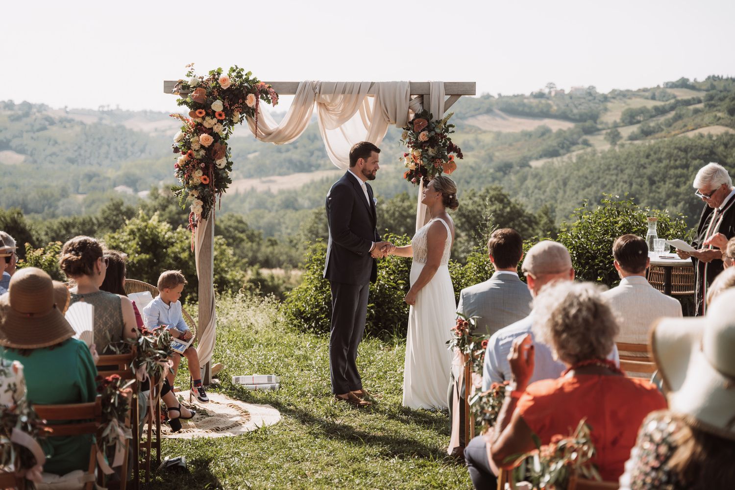 A scenic outdoor wedding ceremony takes place on a hilltop with guests seated under floral-adorned wooden arches.