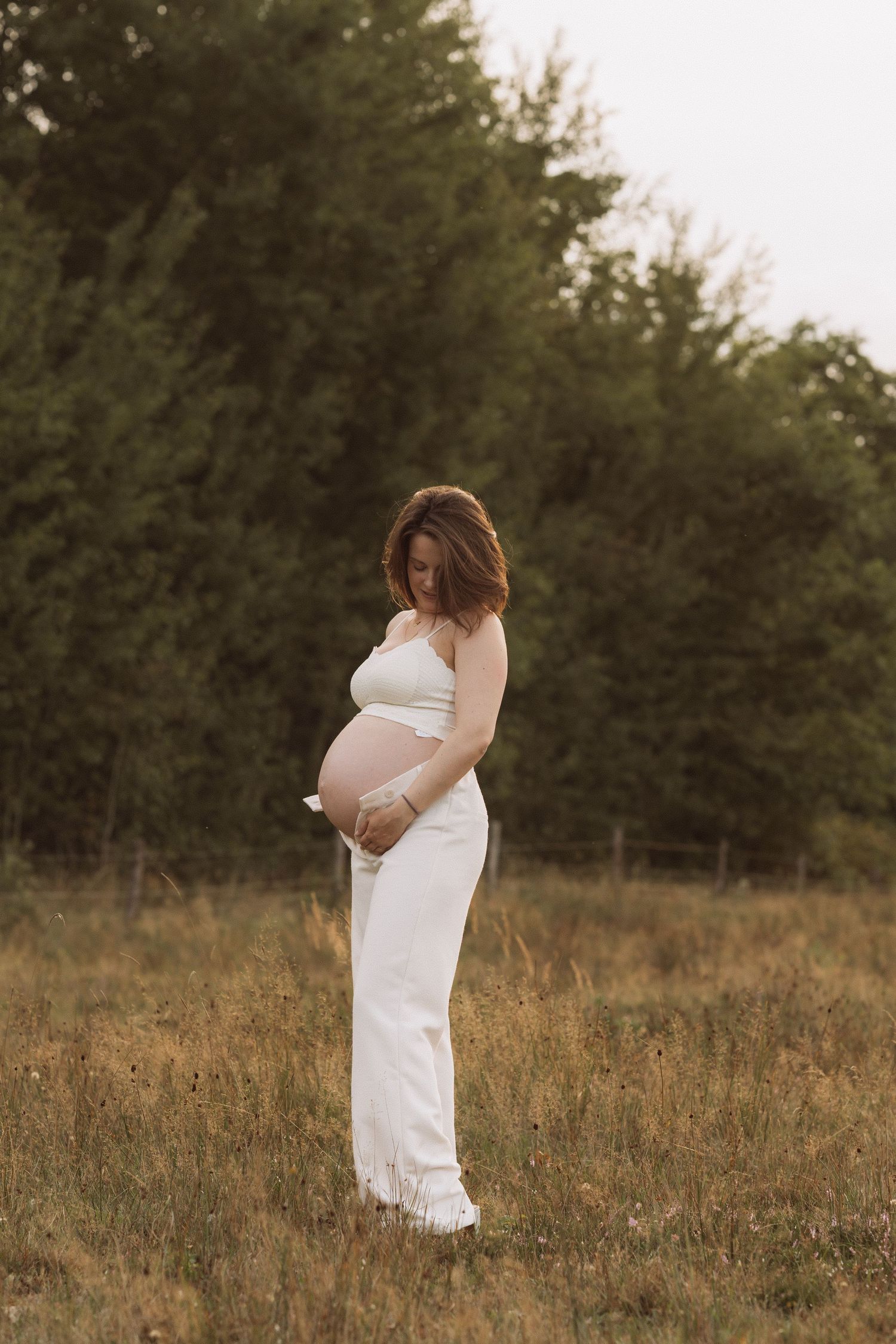 A woman in a white dress stands in a golden field at sunset, cradling her pregnant belly.