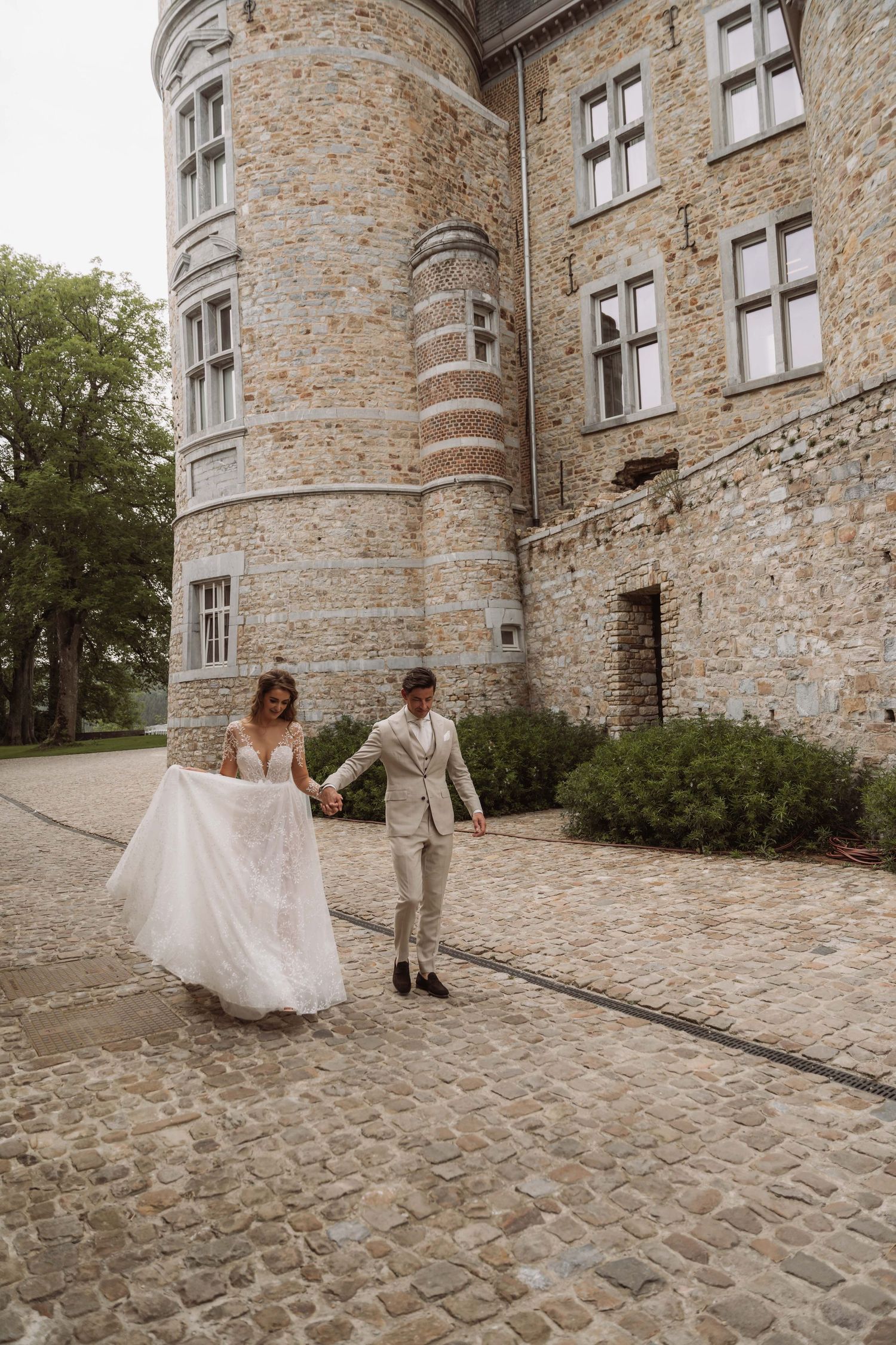 A couple in wedding attire playfully dances and twirls on a cobblestone path beside a historic stone castle wall.