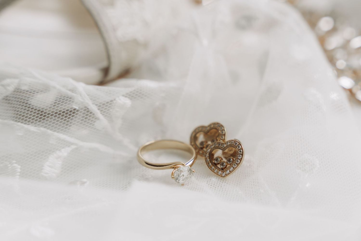 Delicate gold wedding rings and jewelry displayed on white lace fabric with soft bokeh lighting effect in background.