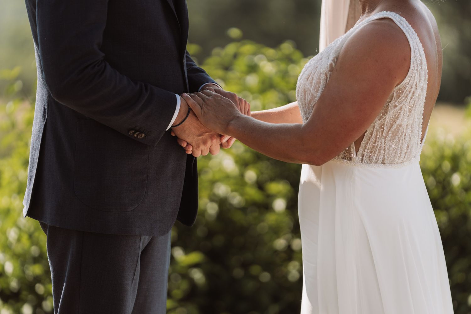A couple holds hands during an outdoor wedding ceremony with sunlit greenery in the background.