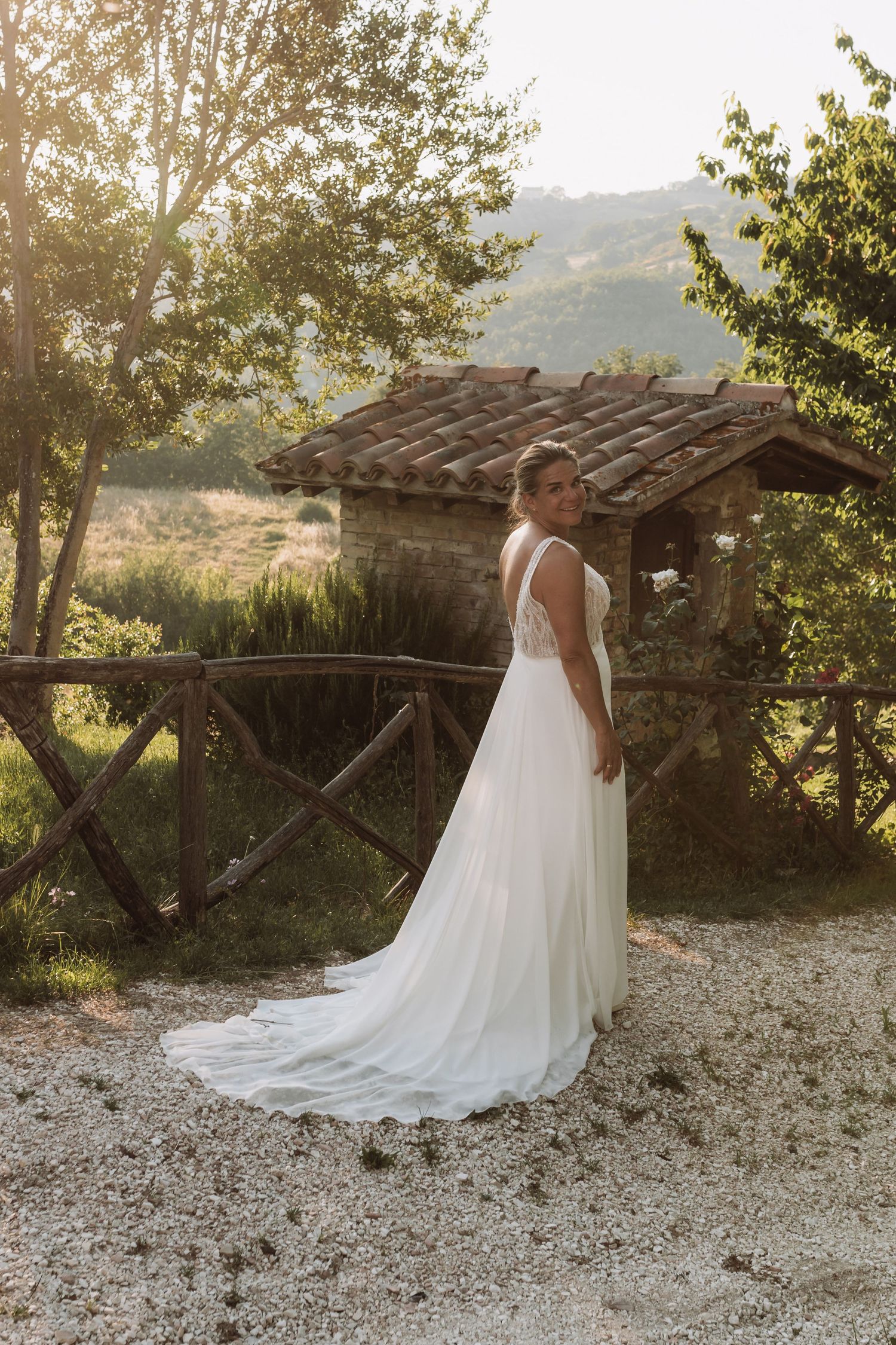 A bride in a flowing white wedding gown poses outdoors against a rustic wooden structure at sunset.