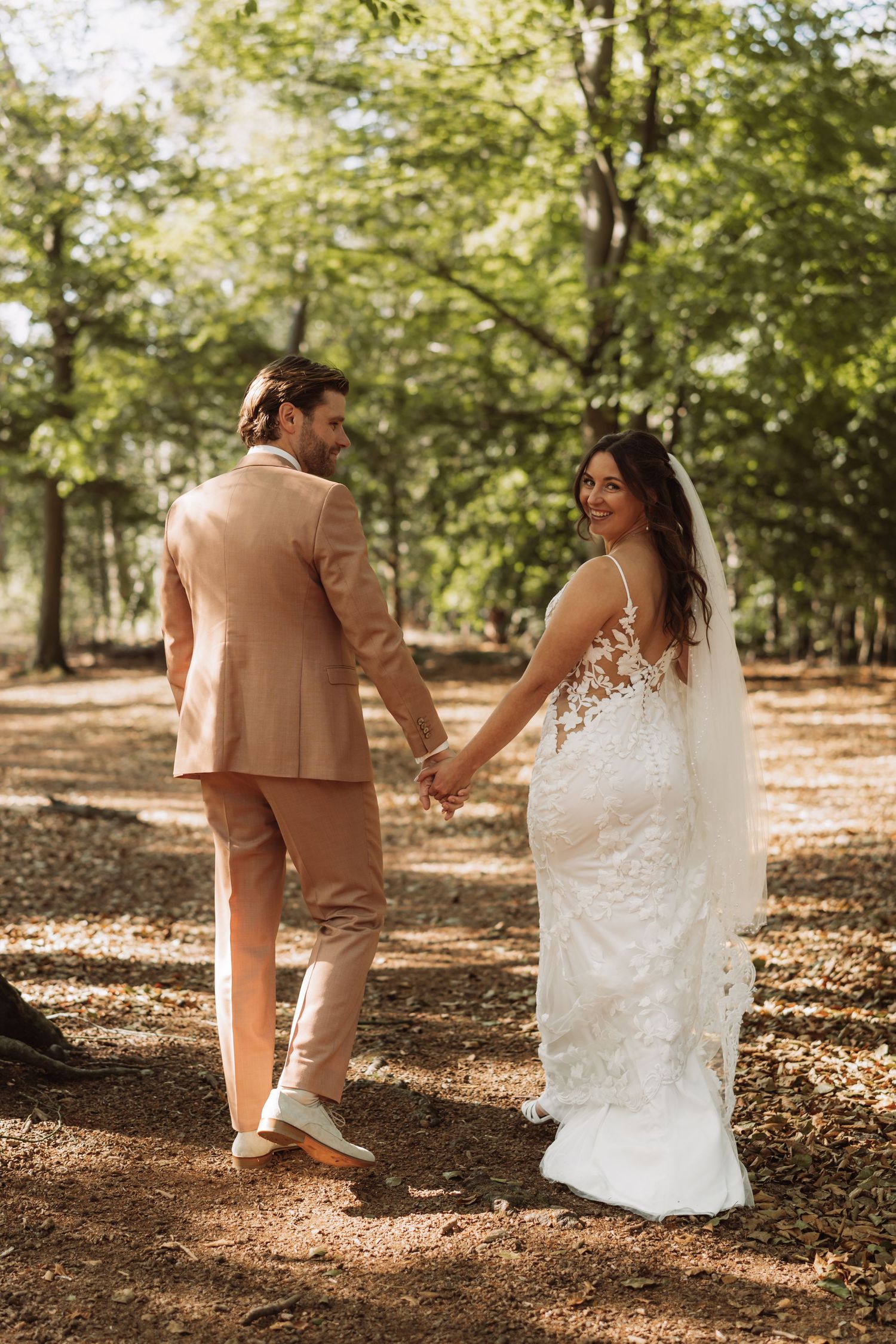 A couple in wedding attire walks together through a sunlit forest with fallen autumn leaves.