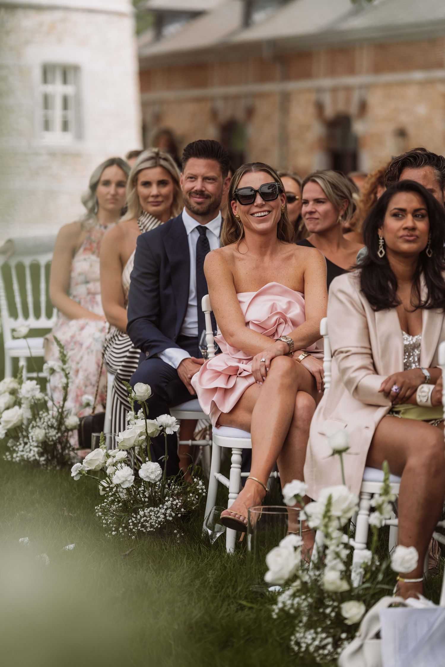 Wedding guests seated outdoors smile and laugh during a ceremony with white floral aisle decorations.