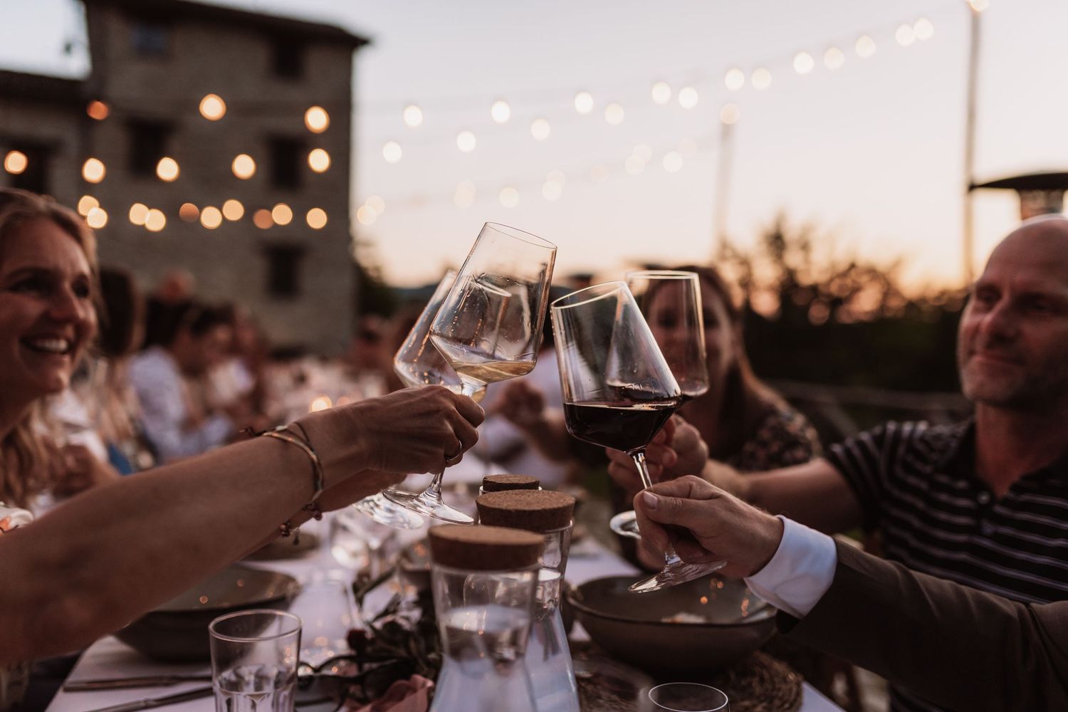 People toasting with wine glasses at an outdoor dinner party with string lights overhead during sunset.