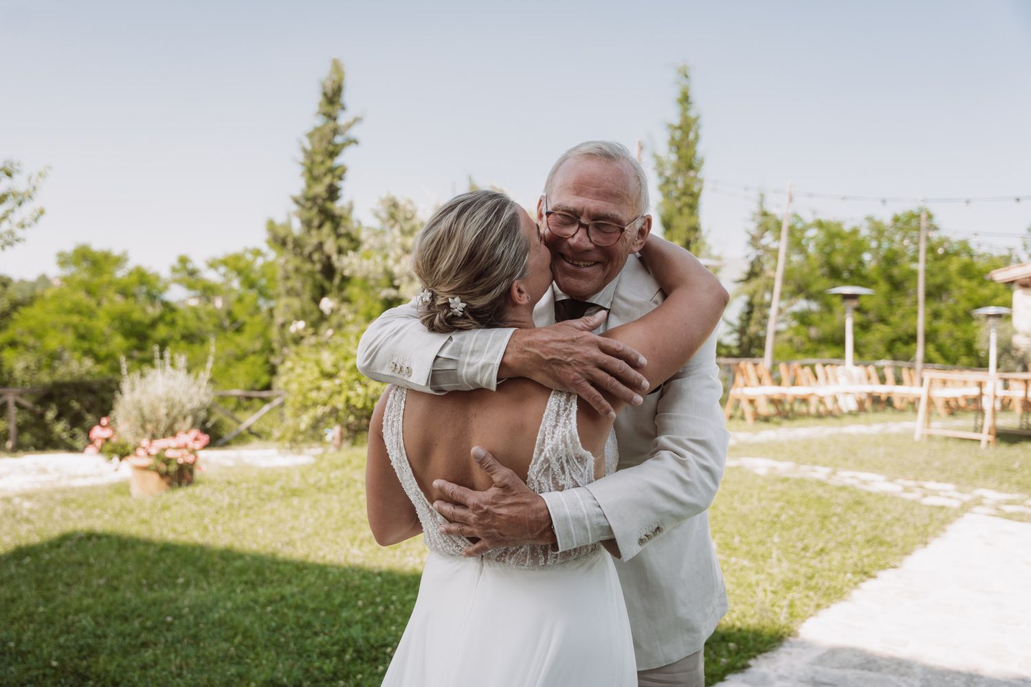 A couple shares a joyful embrace during their outdoor wedding ceremony on a sunny day in a garden setting.