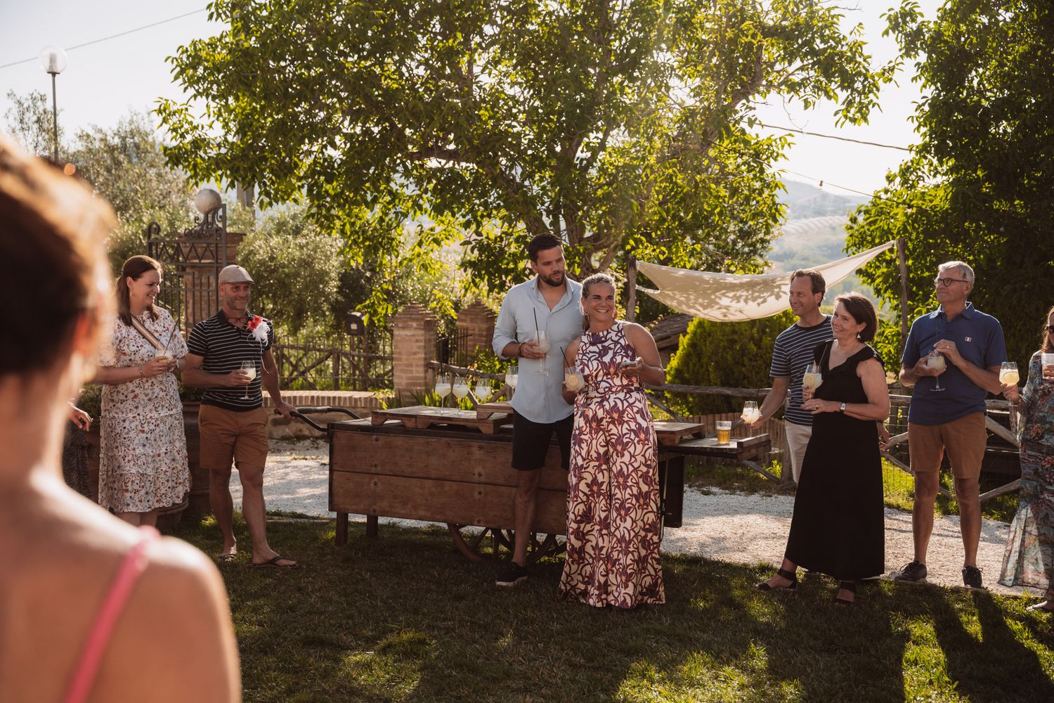 A group gathers around a wooden table at an outdoor summer party in a sunlit backyard garden setting.