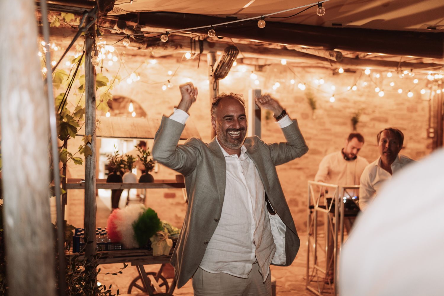 Joyful wedding guests dance and celebrate on an illuminated dance floor under string lights at an indoor reception.