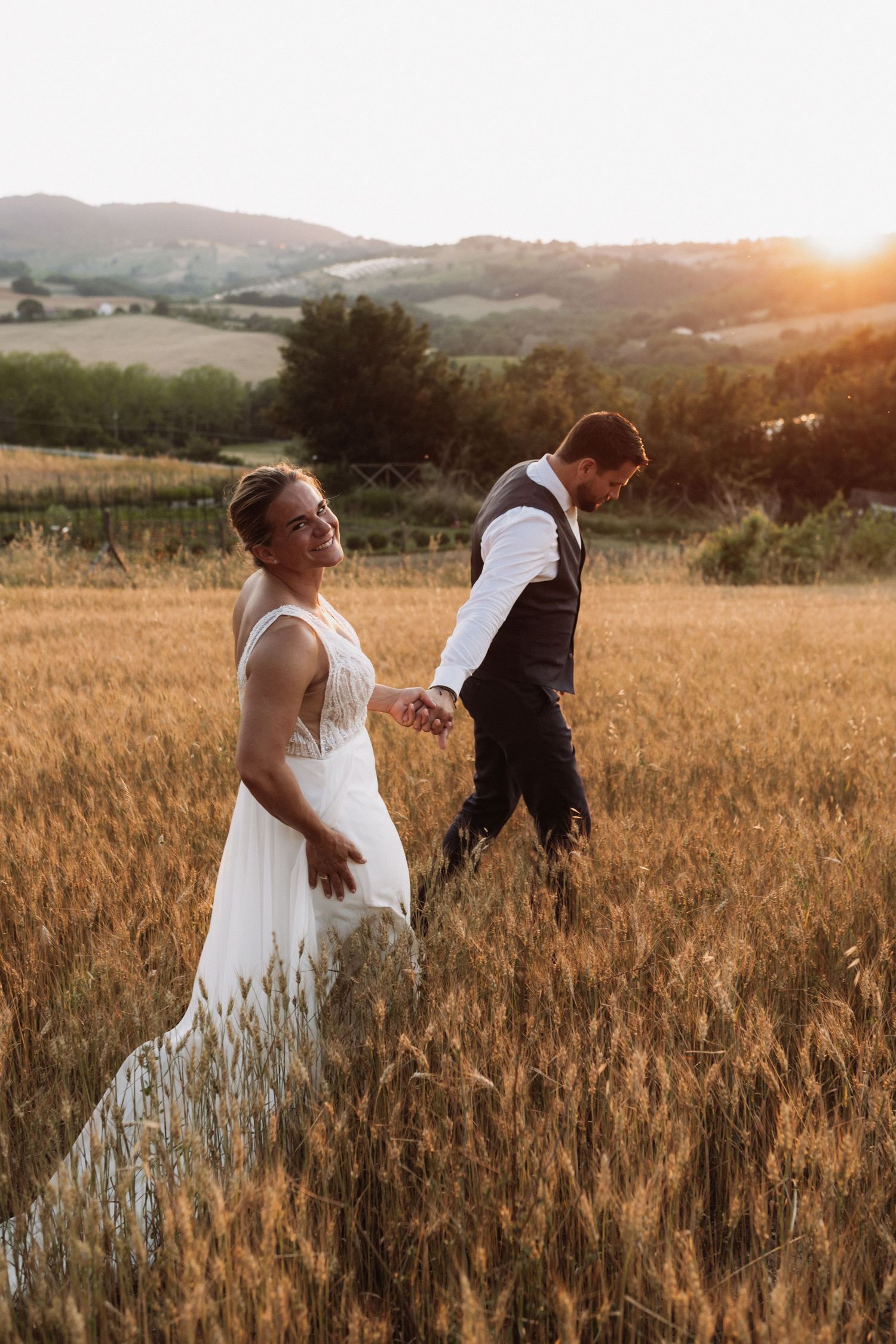 A couple in wedding attire dances playfully in a golden wheat field at sunset in the countryside.