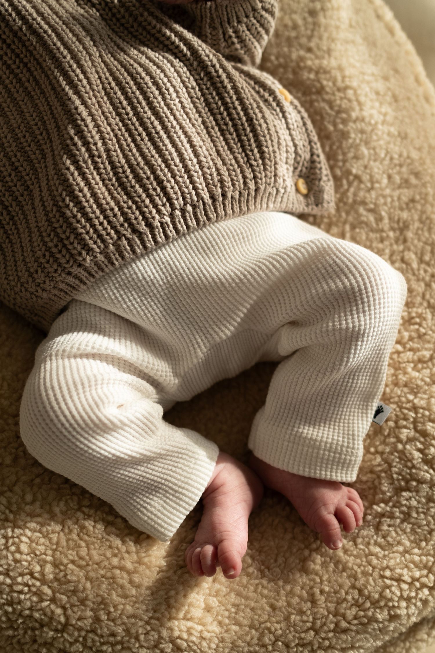 Tiny baby feet peeking out from cozy white knit pants and gray sweater on beige carpet.