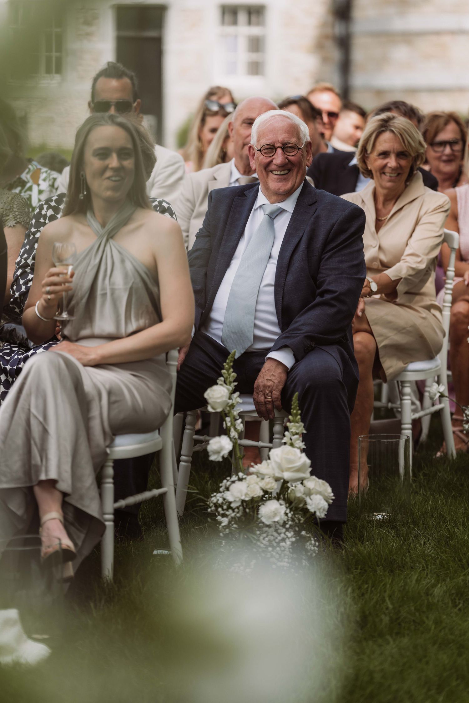 Wedding guests seated outdoors on white chairs laugh and smile during an outdoor ceremony.