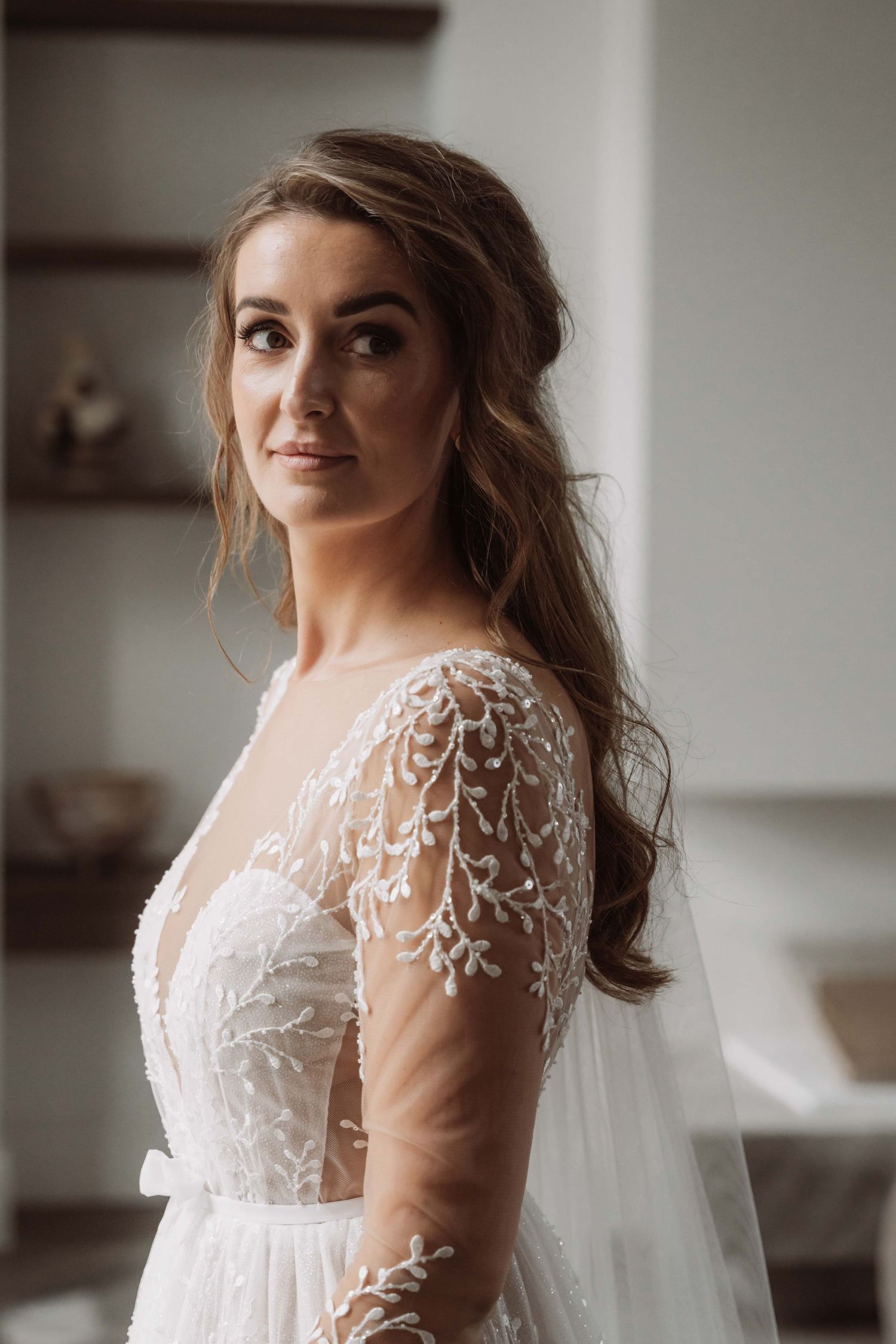 A bride in a delicate white lace wedding dress poses in soft natural light during a bridal photoshoot.