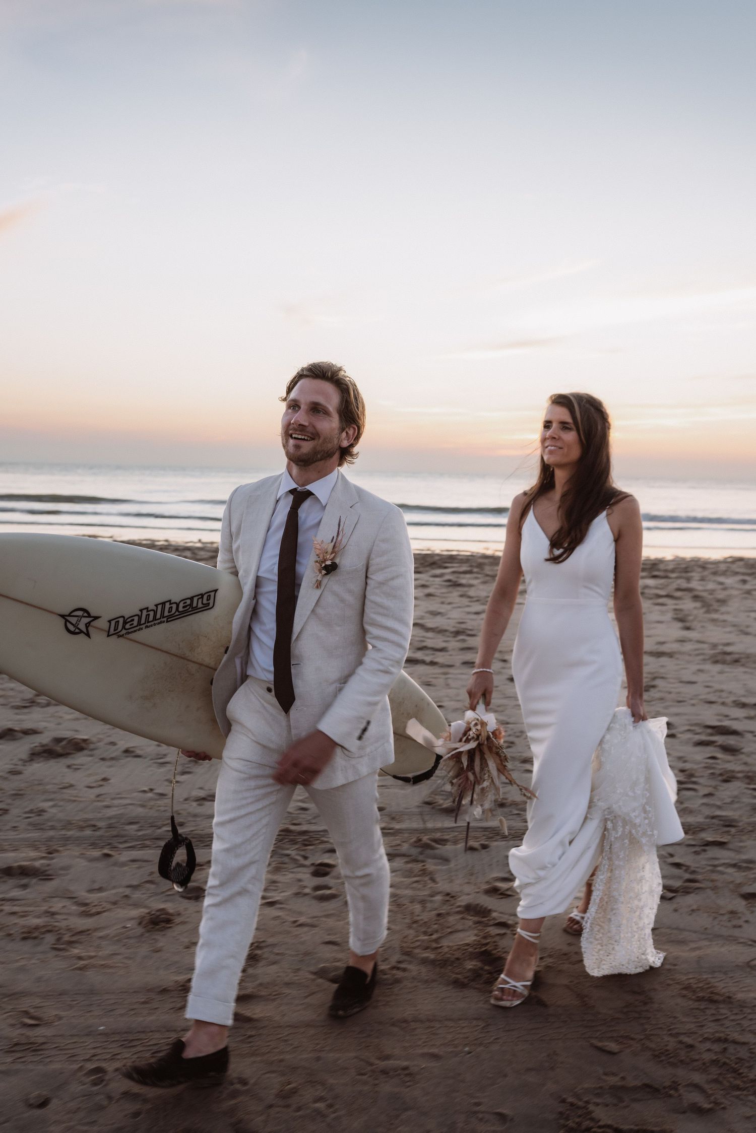 A couple in white wedding attire walks along a beach at sunset carrying a surfboard.