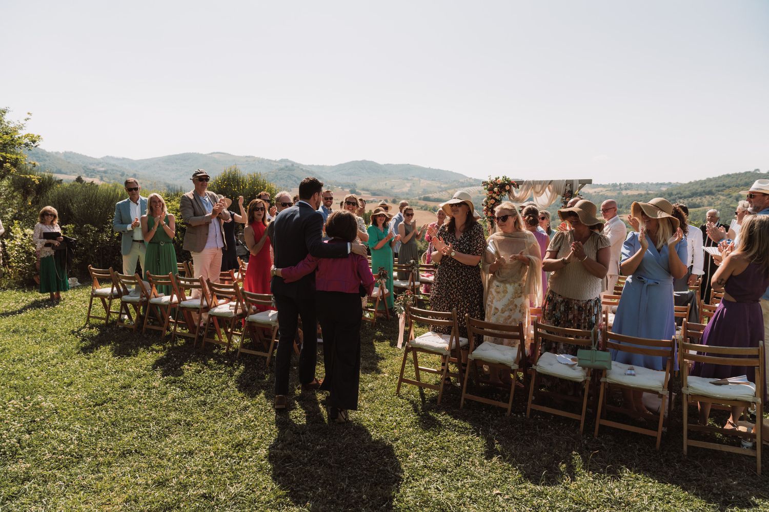 Wedding guests stand during an outdoor ceremony with scenic mountain views on a sunny afternoon.