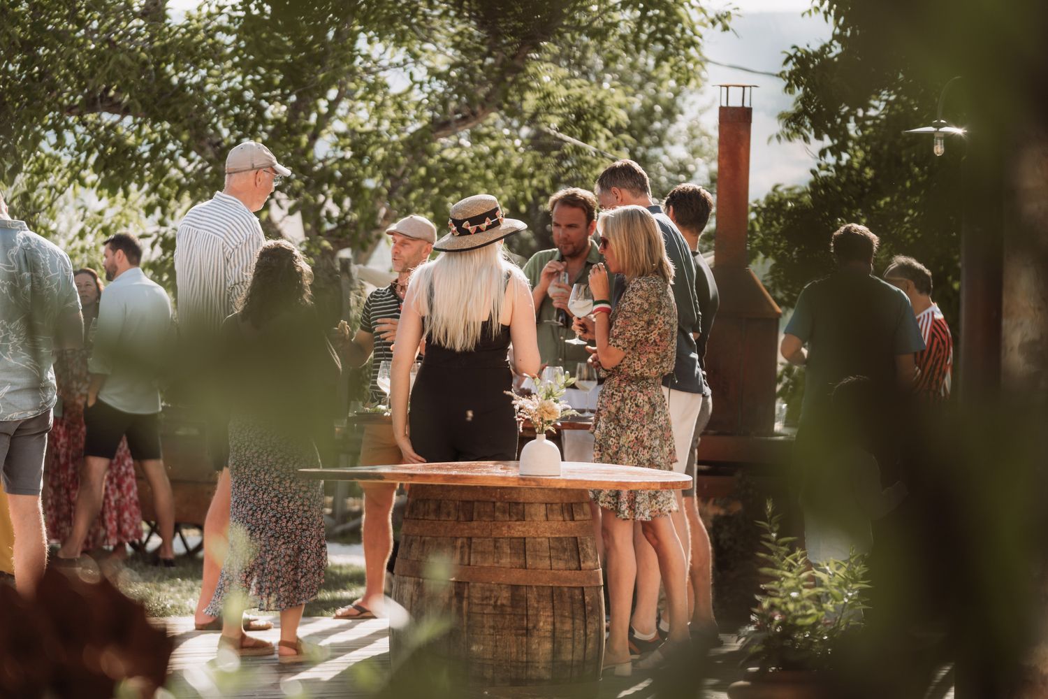 Guests gather around rustic wooden barrels at an outdoor summer party in a garden setting.