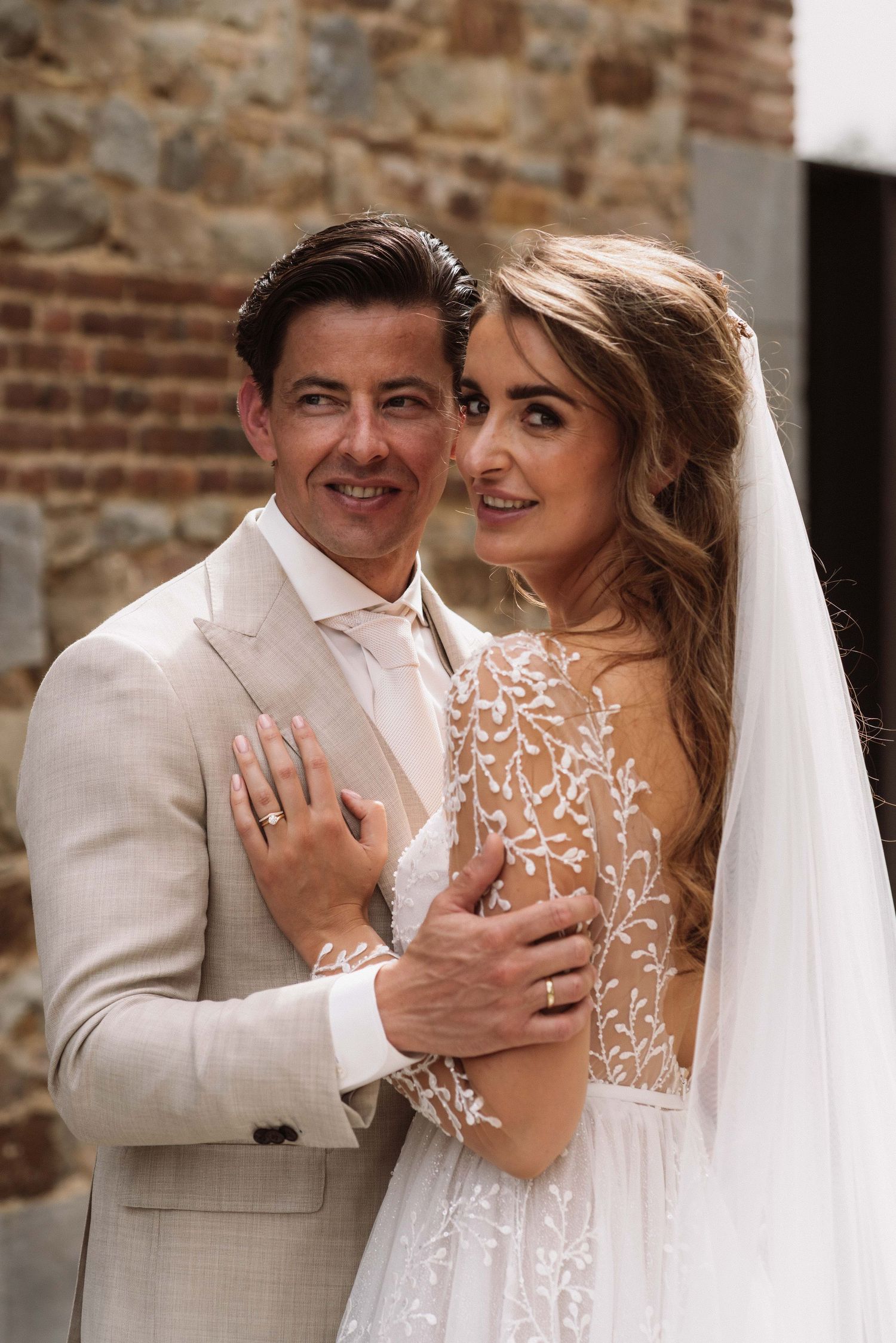 Intimate wedding portraits against rustic brick wall showing couple in beige suit and delicate lace gown.