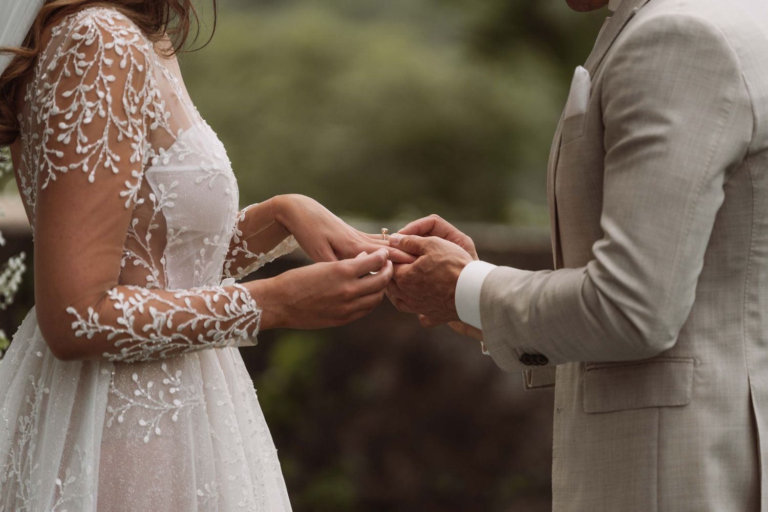 A couple exchanges rings during an outdoor wedding ceremony with lace details and natural lighting.