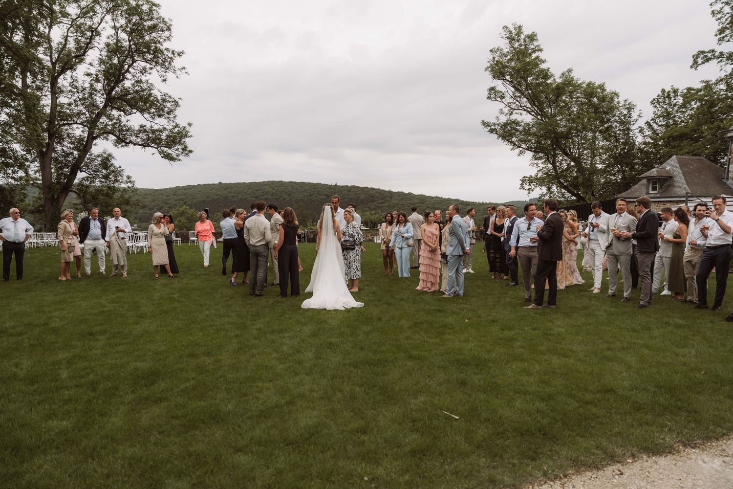 Wedding ceremony guests gather on a grassy hillside overlooking scenic mountain views on a cloudy day.