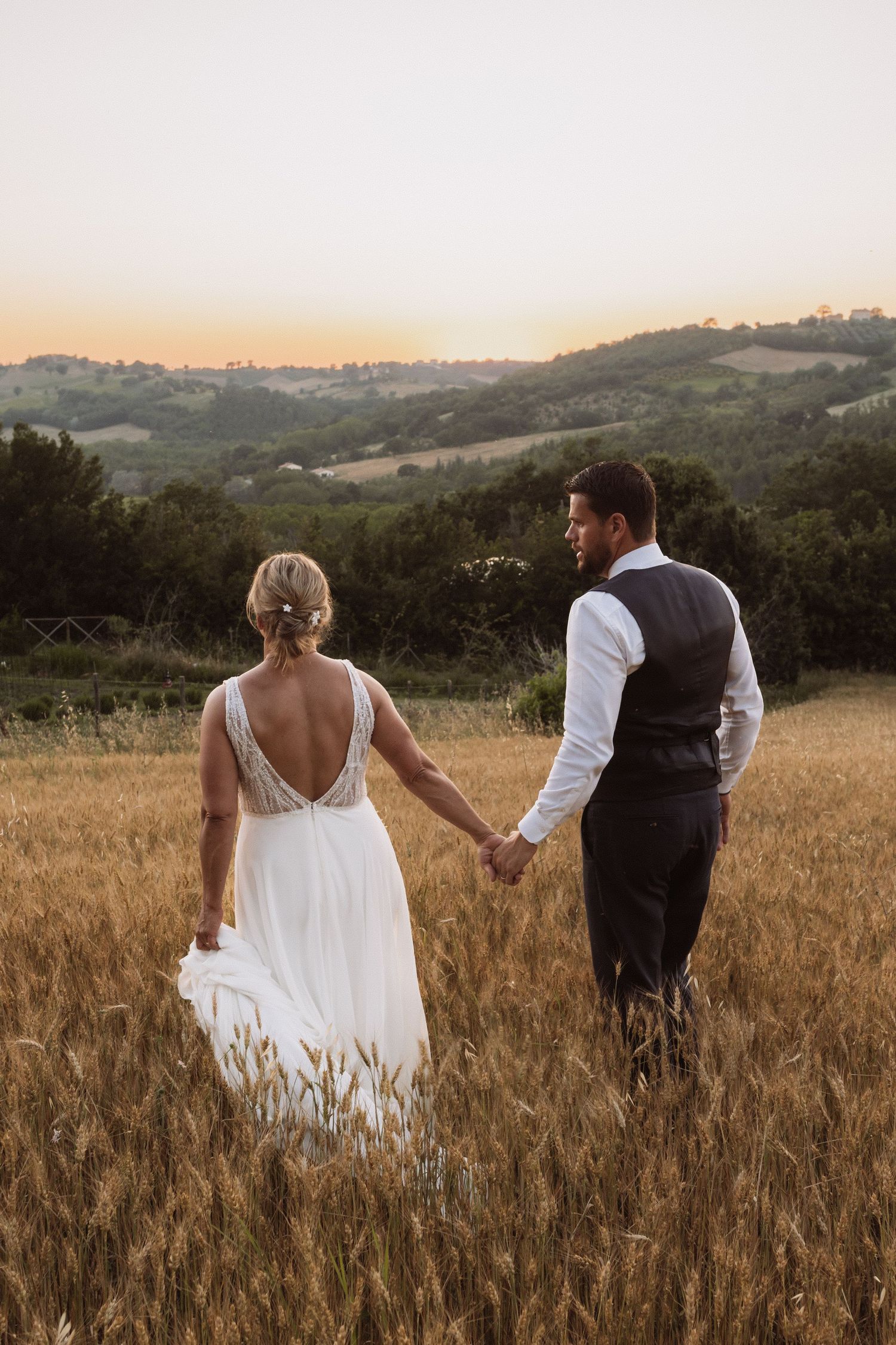 A couple holding hands while walking through a golden wheat field at sunset.