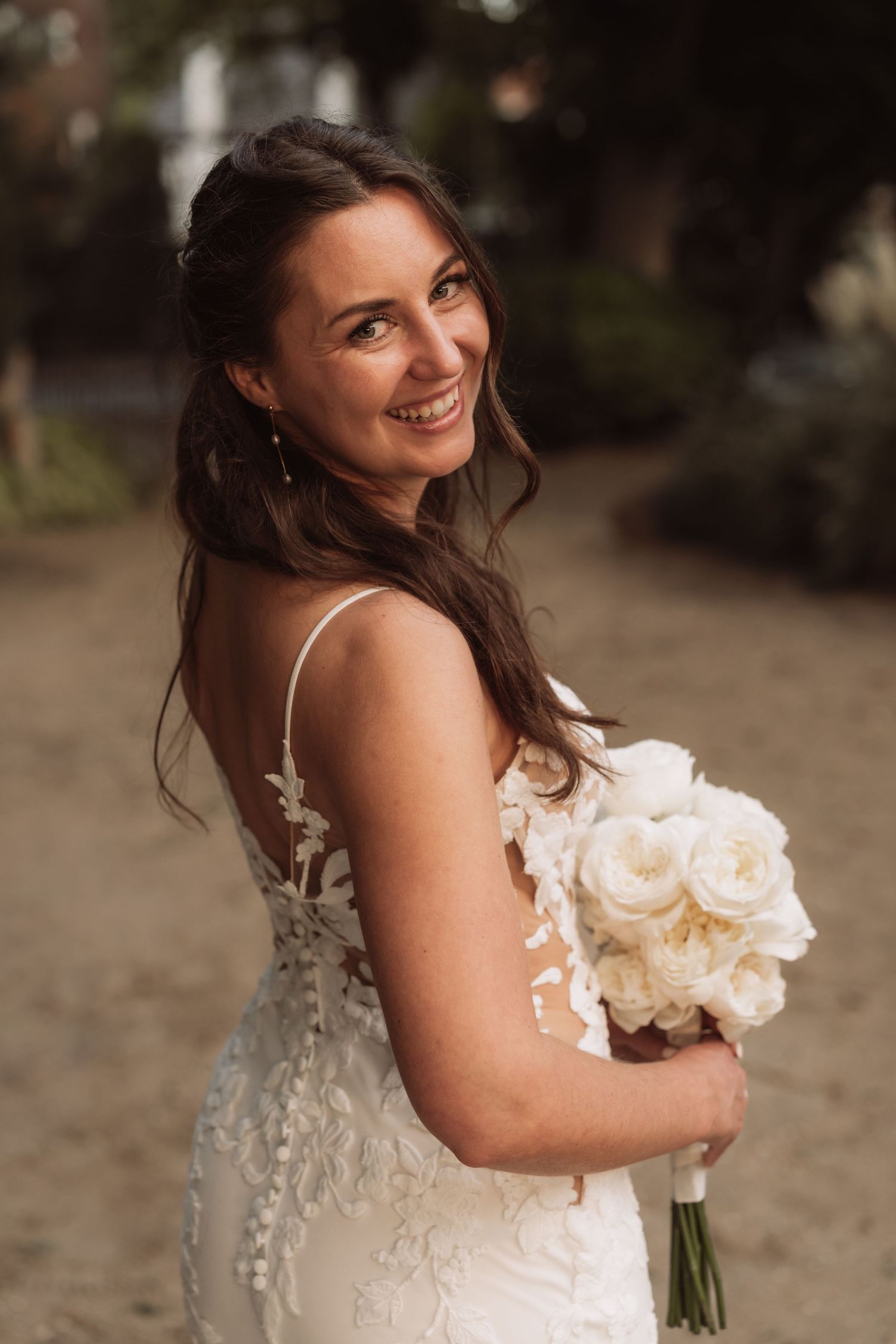 A bride in a white lace wedding dress holds a bouquet of white roses while smiling and looking over her shoulder.