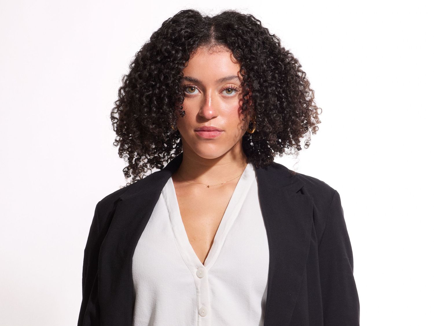 Young woman with curly hair wearing a black blazer and white blouse poses against a white background. Photo taken in Salem, OR