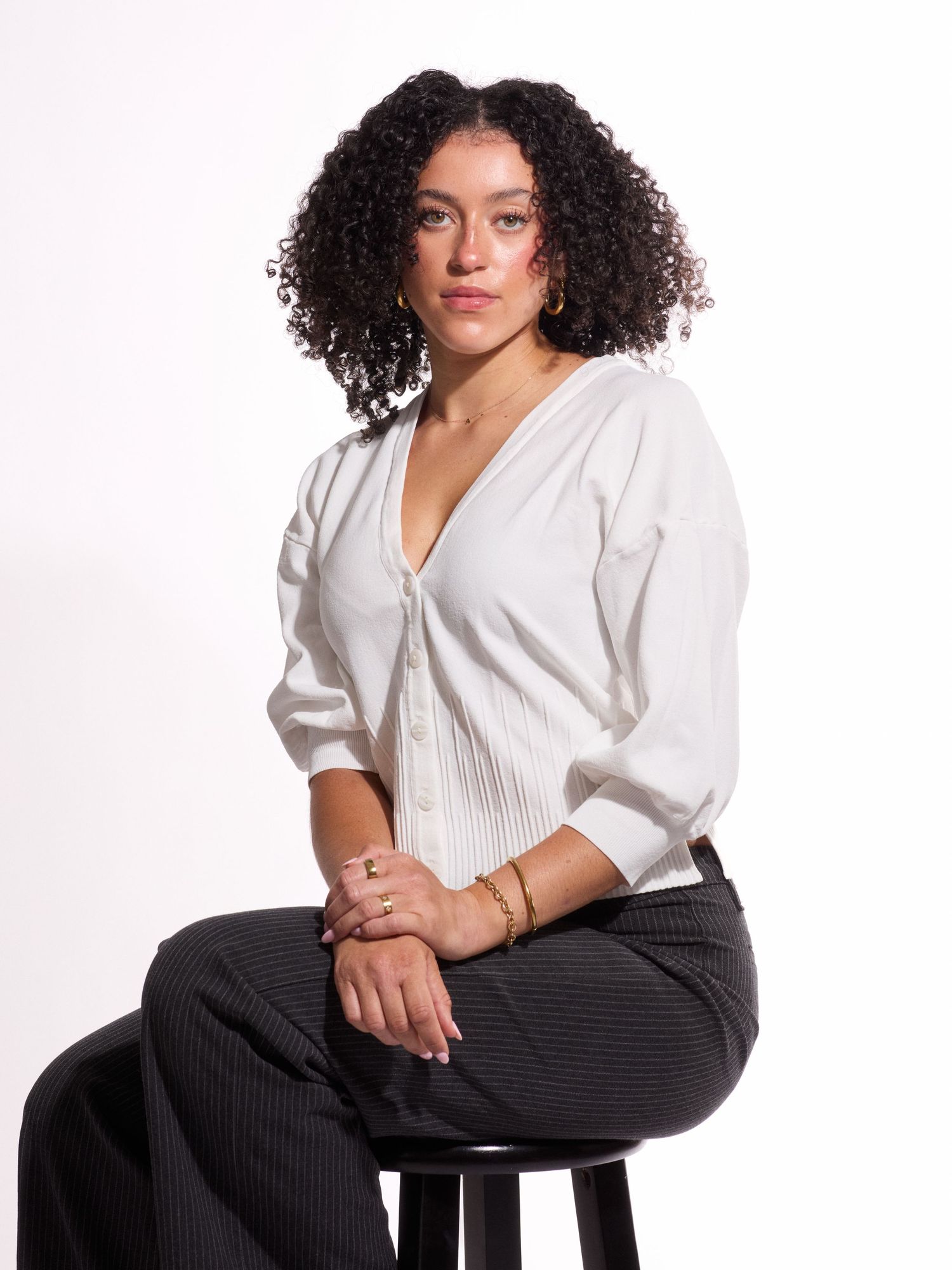 Woman with curly hair sits on stool wearing white blouse and black pinstripe trousers against white background.