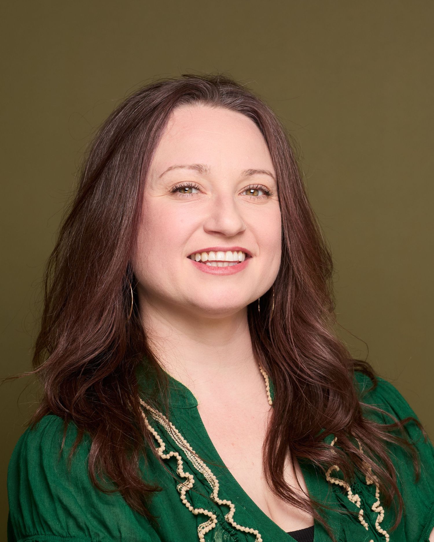 Smiling woman with long brown hair wearing a green ruffled blouse and pearl necklace against an olive background. Photo taken in springfield