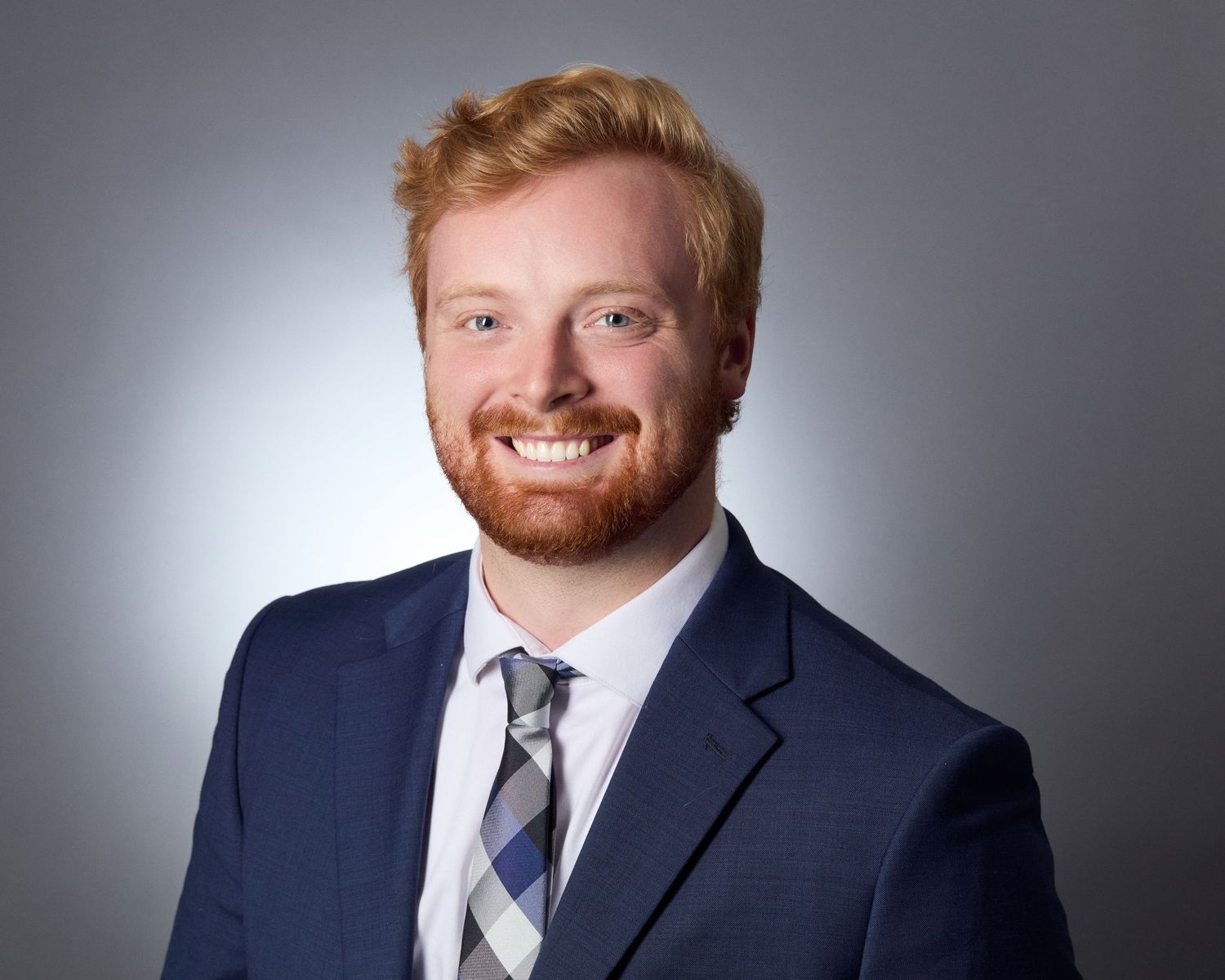 Professional corporate headshot of a smiling young man wearing a navy blue suit and tie. Photo taken on site in Eugene, OR