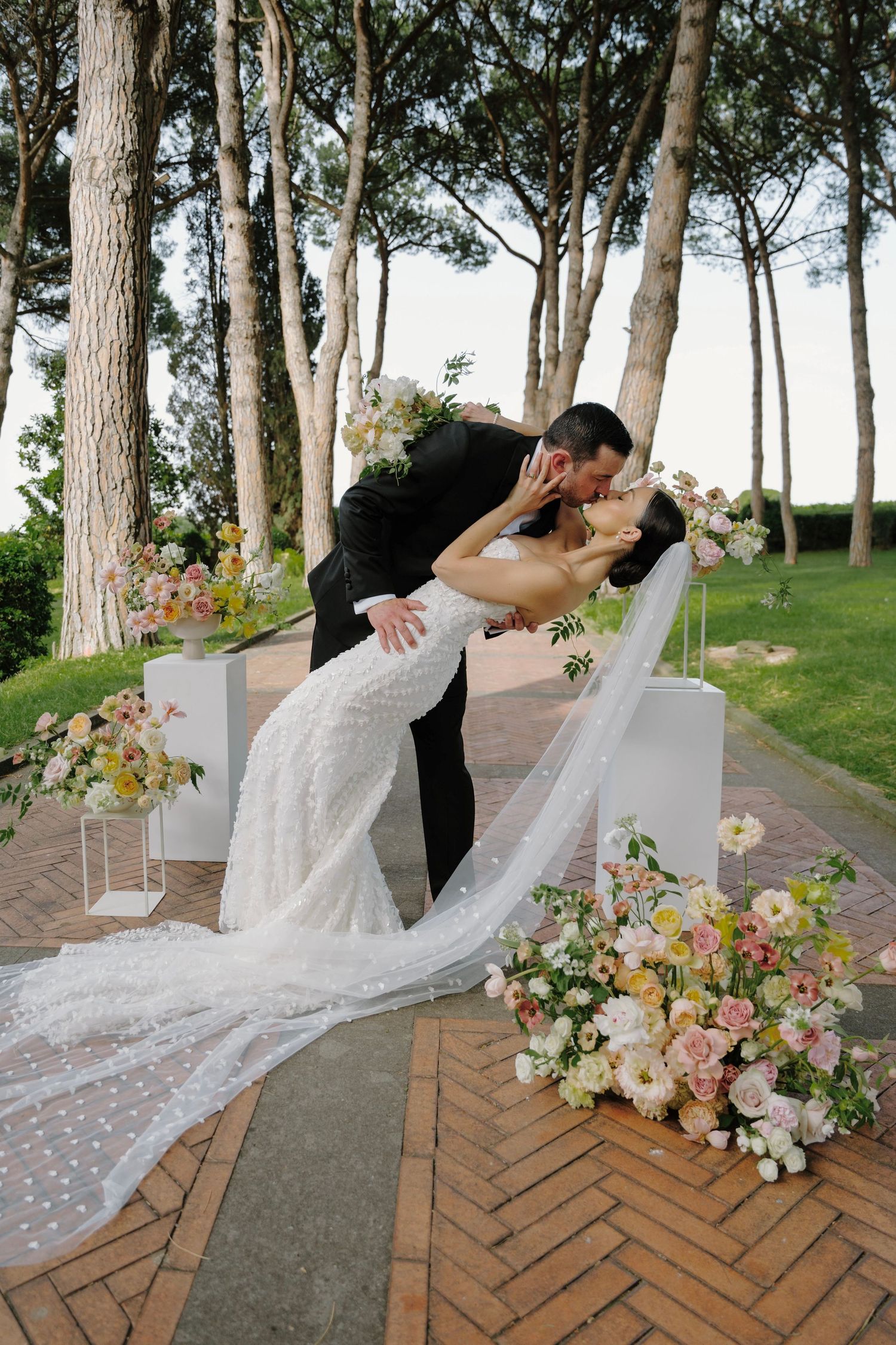 Romantic dip pose with flowing veil and floral arrangements on brick pathway.