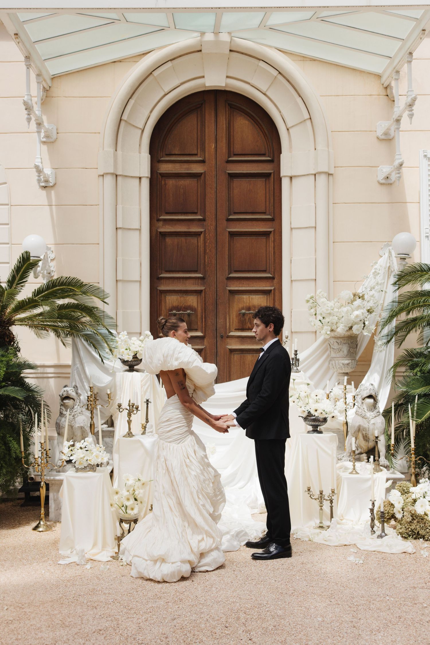 Wedding couple poses in front of grand wooden doors with white floral arrangements and palm decor.